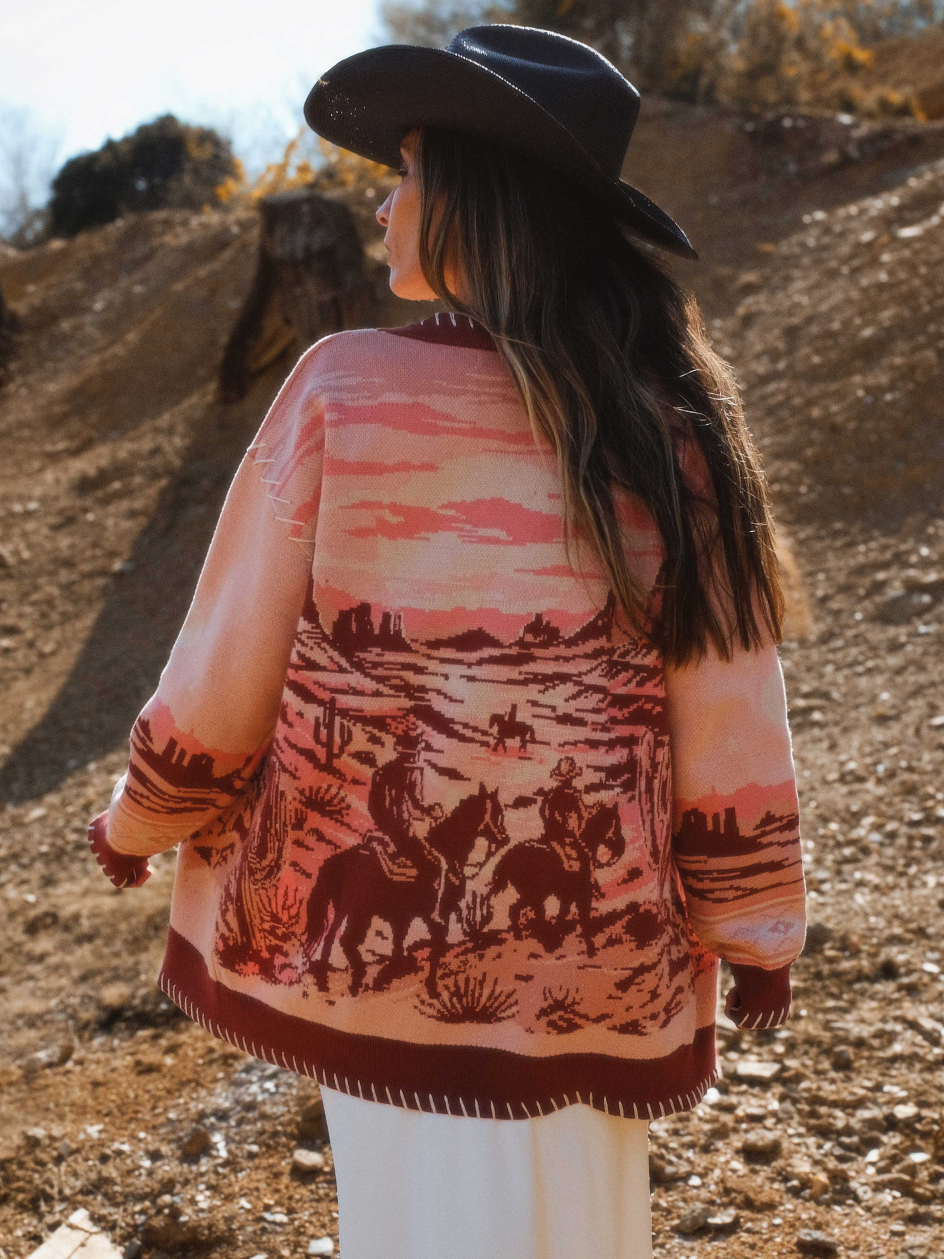Back view of a person wearing a pink knit cardigan with a western desert landscape knit pattern featuring horses and riders.