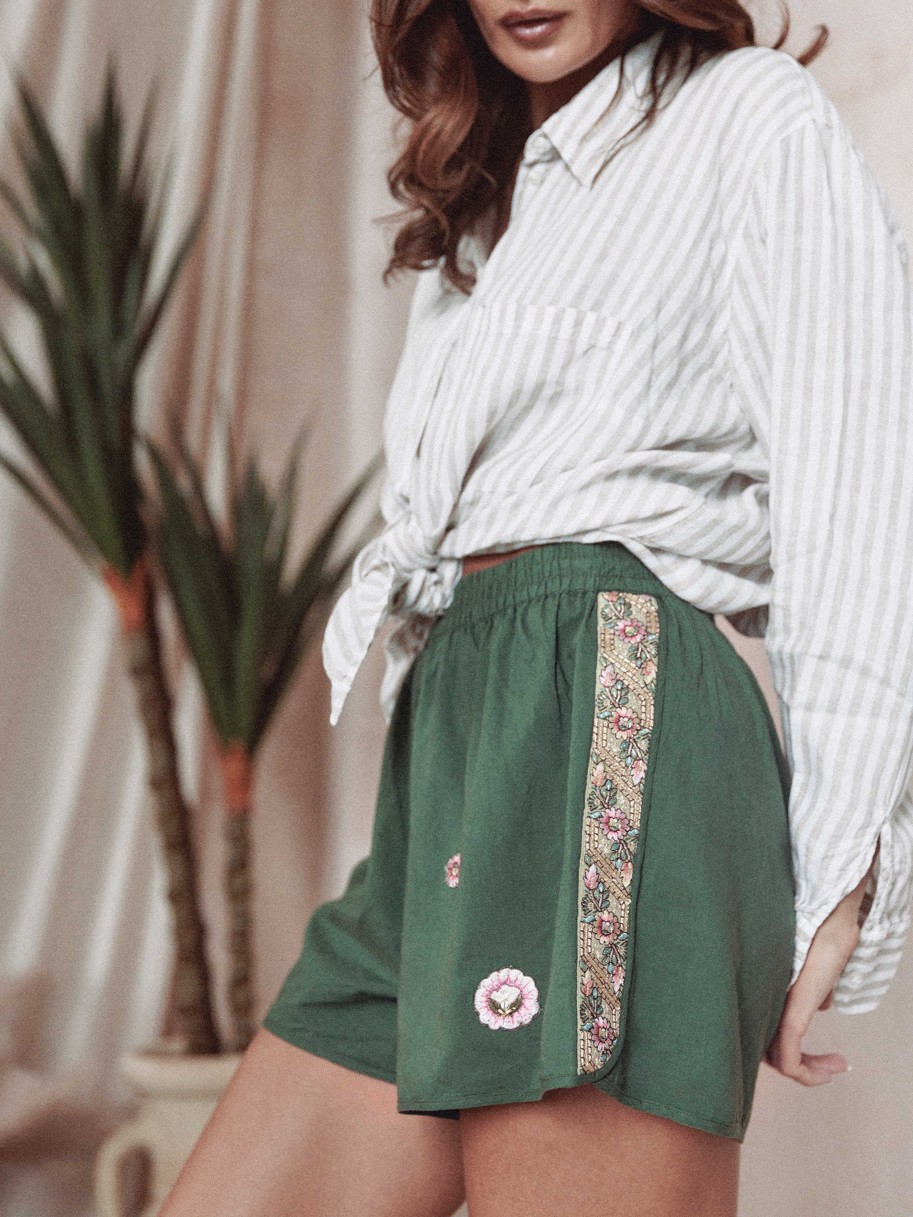Person wearing green shorts with floral embroidered details and decorative side trim, paired with a white striped button-up shirt.