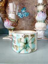 A decorative ceramic candle holder in the center, featuring a pastel blue and cream striped design with a green starburst motif, gold floral accents, and the words 'MERRY CHRIST'. It is styled on a light blue surface with vintage bird candlesticks in the background.