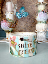 A decorative ceramic candle holder with the words 'SHINE BRIGHT' in gold lettering, decorated with a pink flower, green leaves, and gold stars on a light blue background. It is styled on a tabletop with bird figurines and candlesticks in the background.