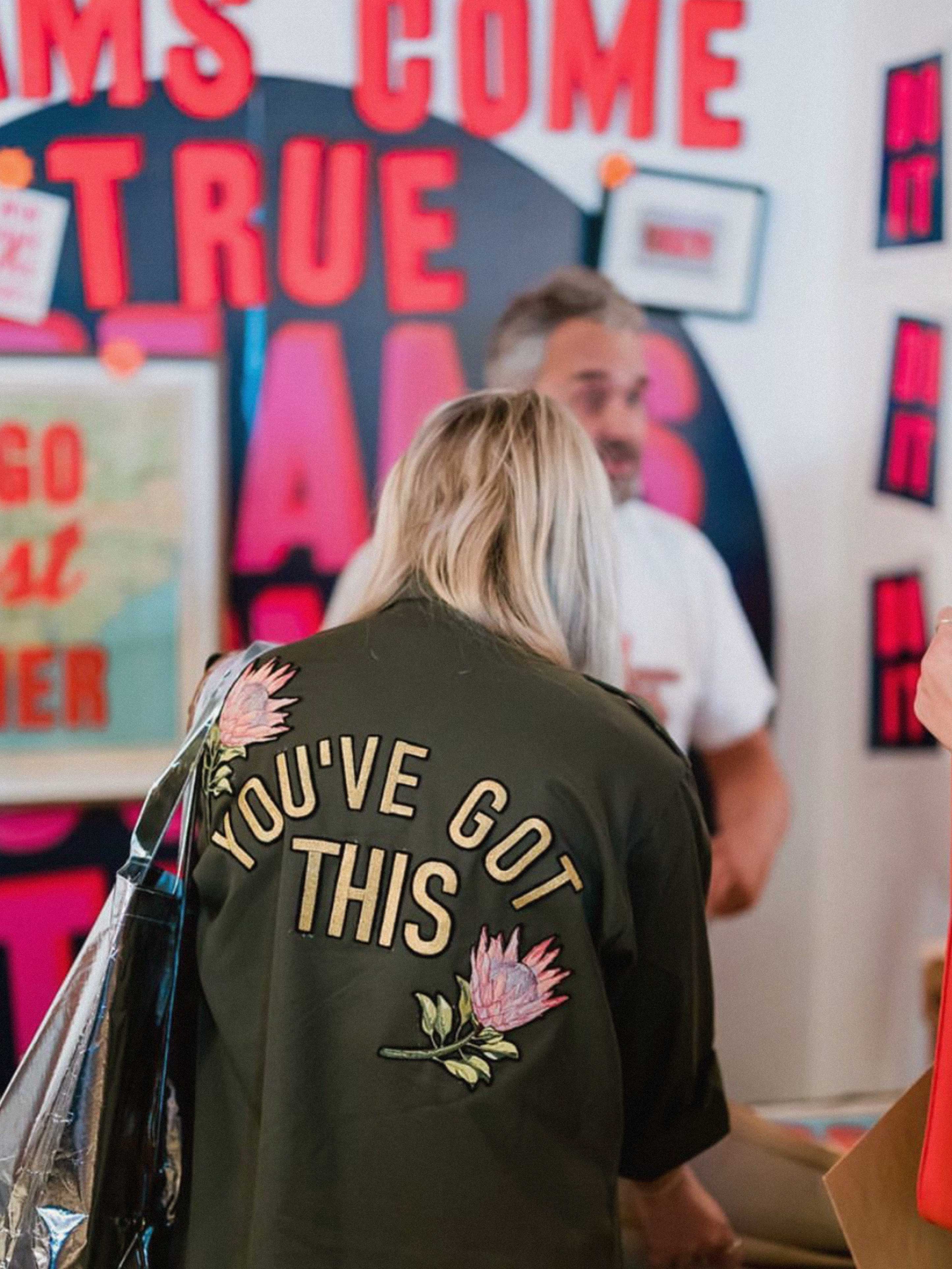 Back view of a person wearing an olive green military jacket with "YOU'VE GOT THIS" embroidery and pink protea flowers in a boutique.