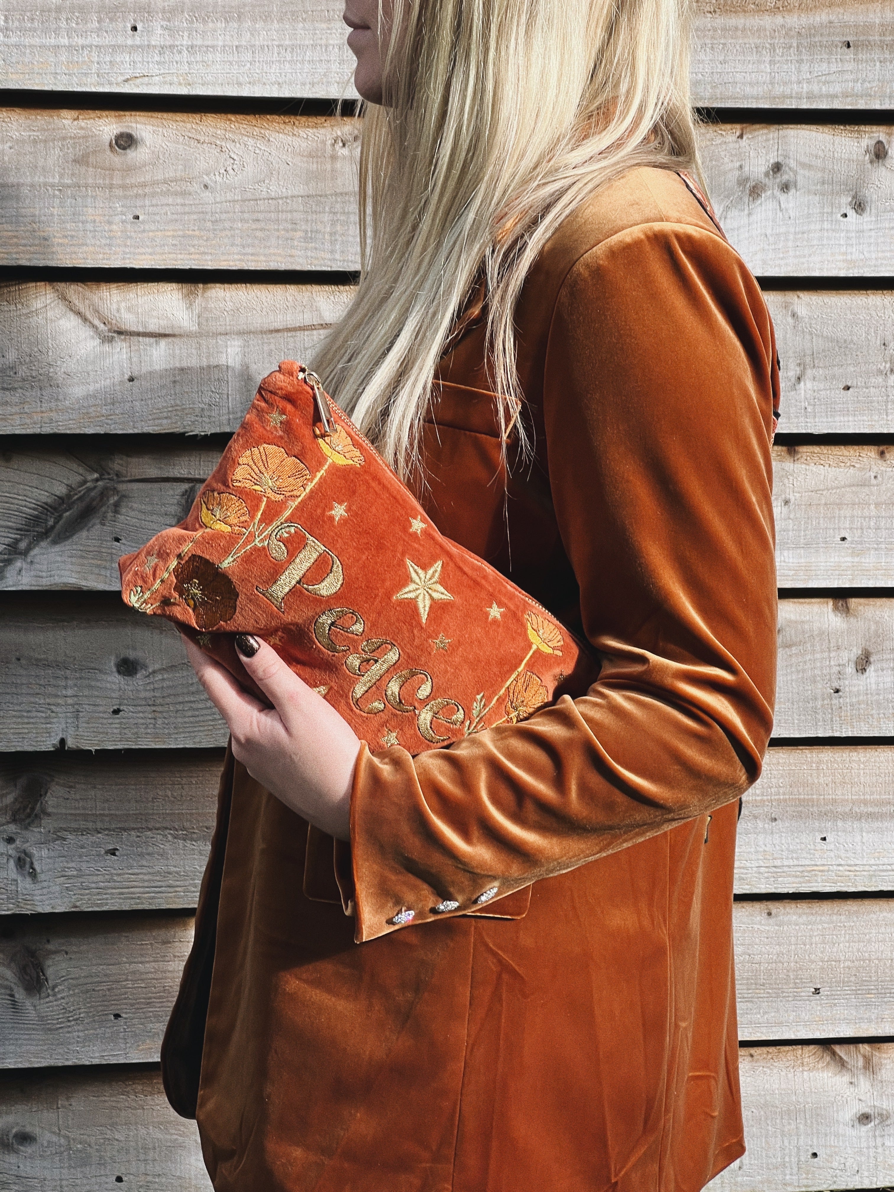 Person holding a rich rust velvet zip pouch embroidered with the word "Peace," wildflowers, and stars.