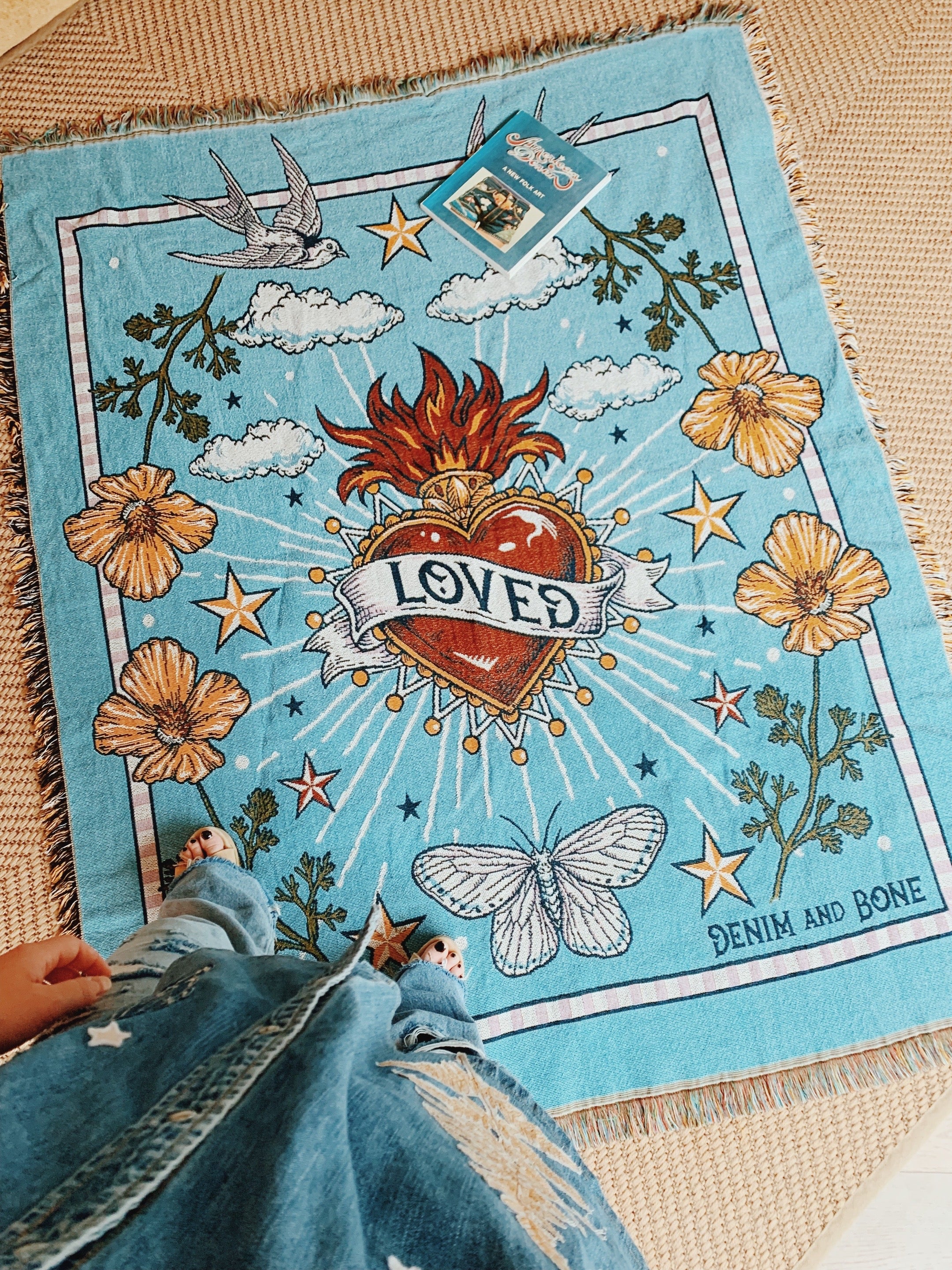 Person holding a light blue "Loved" woven throw blanket featuring a heart, swallows, wildflowers, and moths.