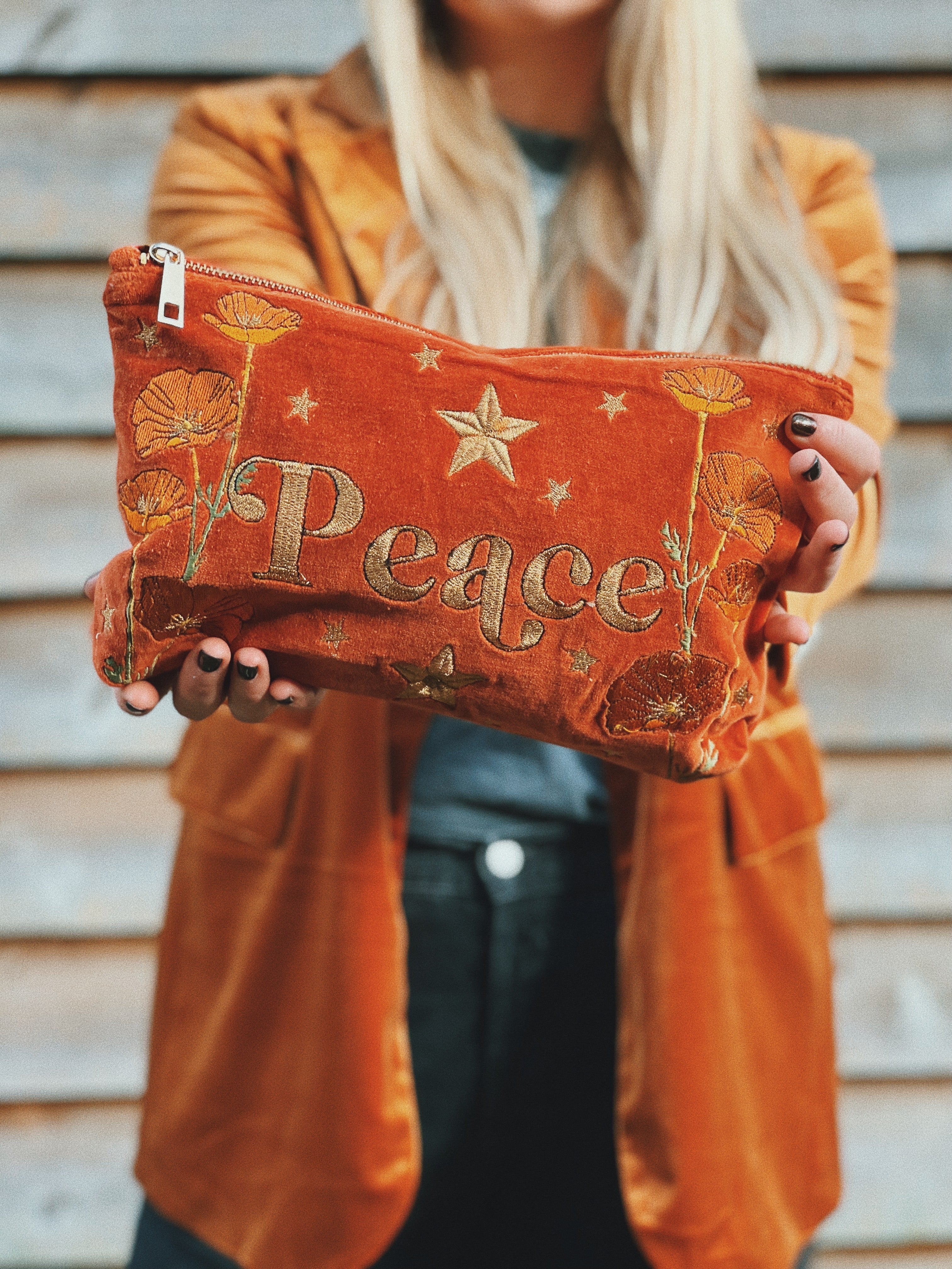 Person holding a rich rust velvet zip pouch embroidered with the word "Peace," wildflowers, and stars.