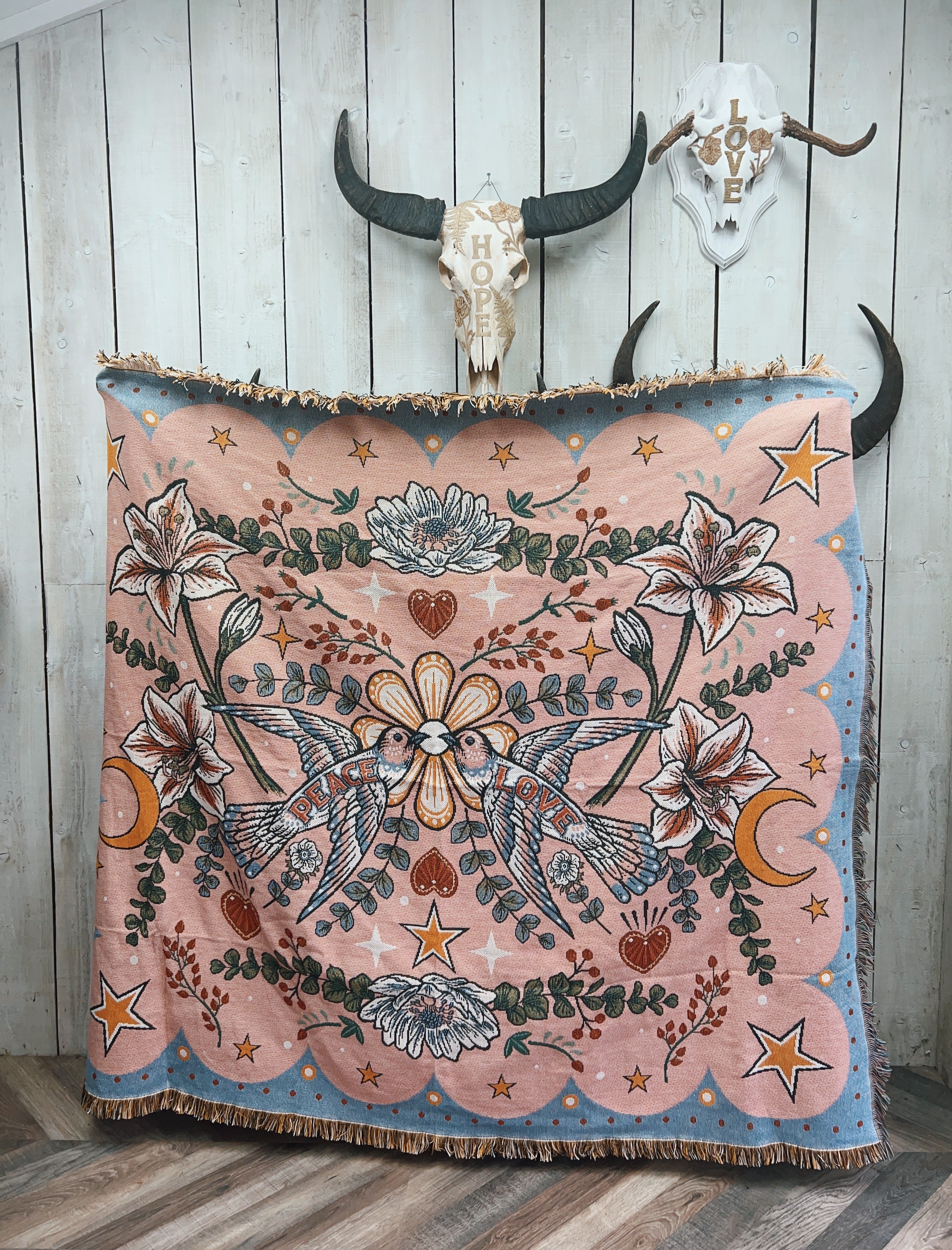 Pink woven throw blanket with hummingbirds, lilies, stars, and moons displayed hanging indoors.