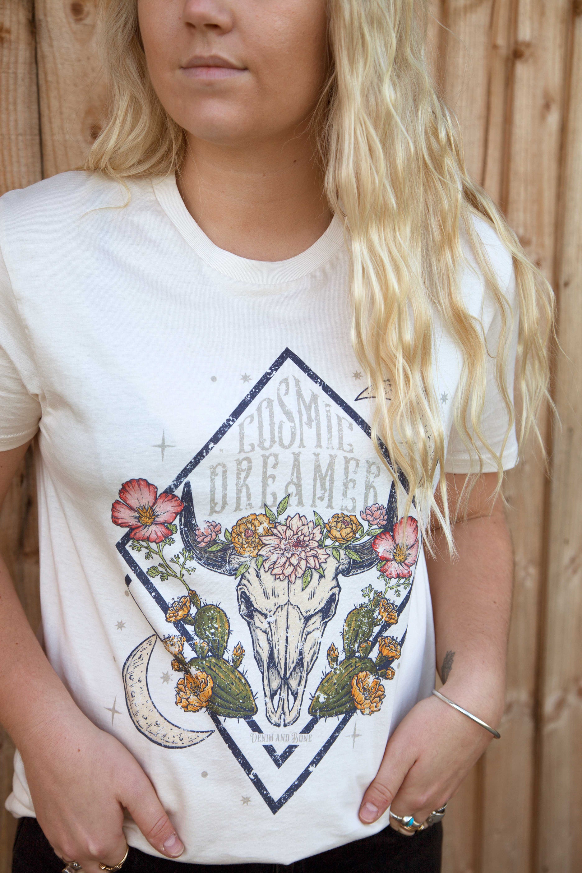 Person wearing a white graphic T-shirt featuring a floral longhorn skull design with crescent moons, desert cacti, and "Cosmic Dreamer" text.