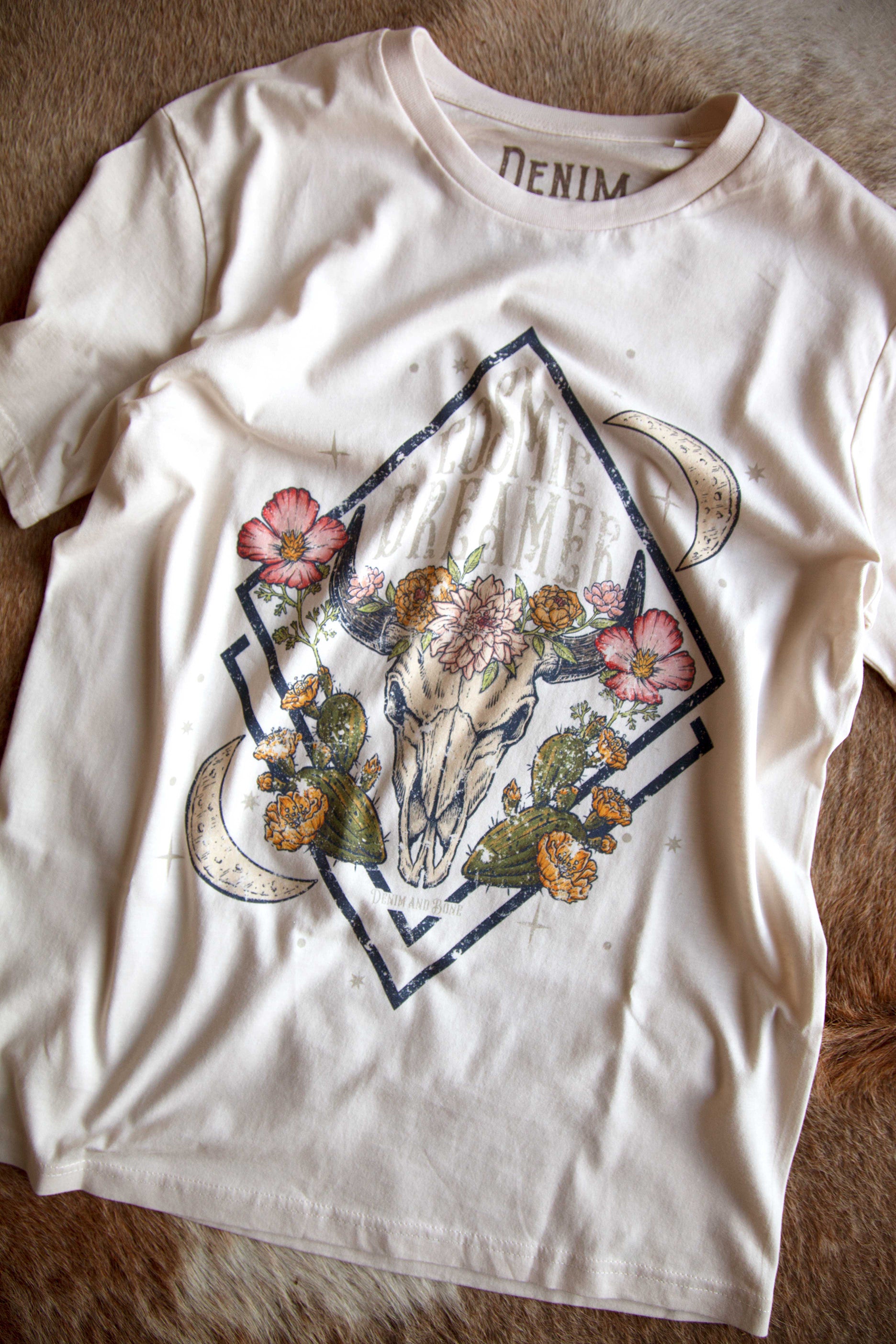 White graphic T-shirt featuring a floral longhorn skull design with crescent moons, desert cacti, and "Cosmic Dreamer" text displayed on a cowhide background.