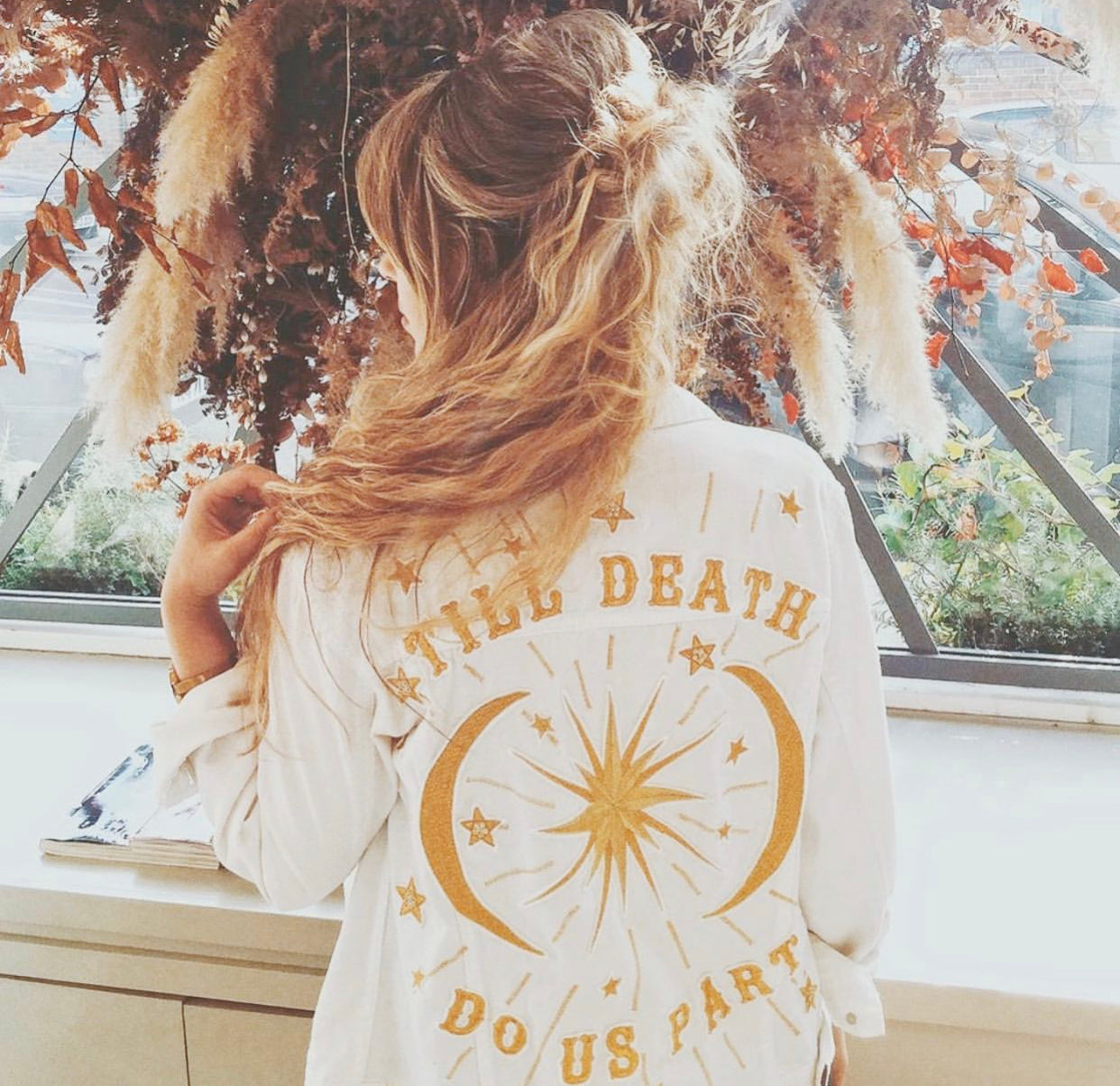 Bride wearing a white embroidered "Till Death Do Us Part" jacket with a celestial design in a floral setting.