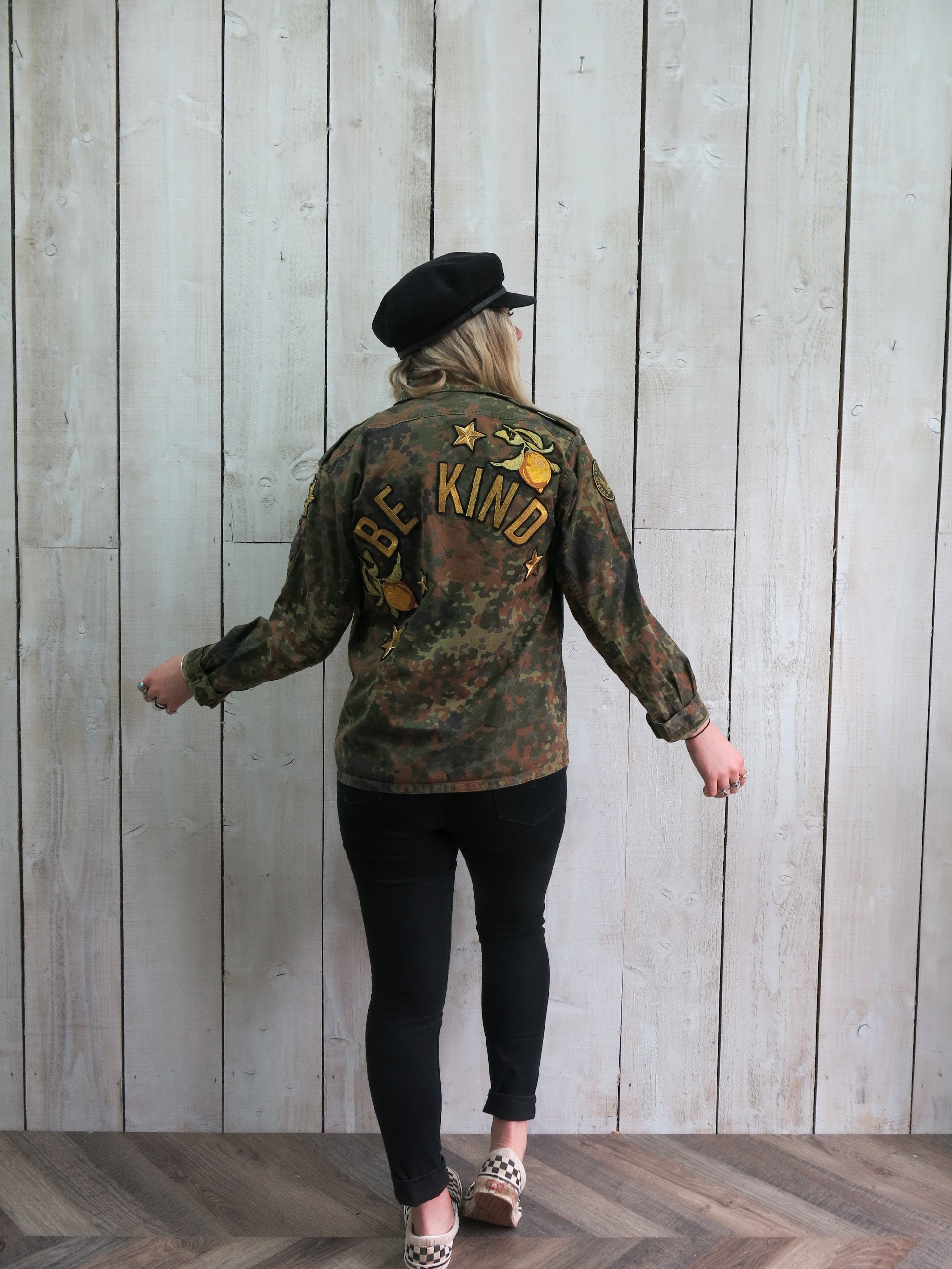 Person wearing a camouflage military jacket with "BE KIND" embroidered on the back, accented by lemons and star embroidery.