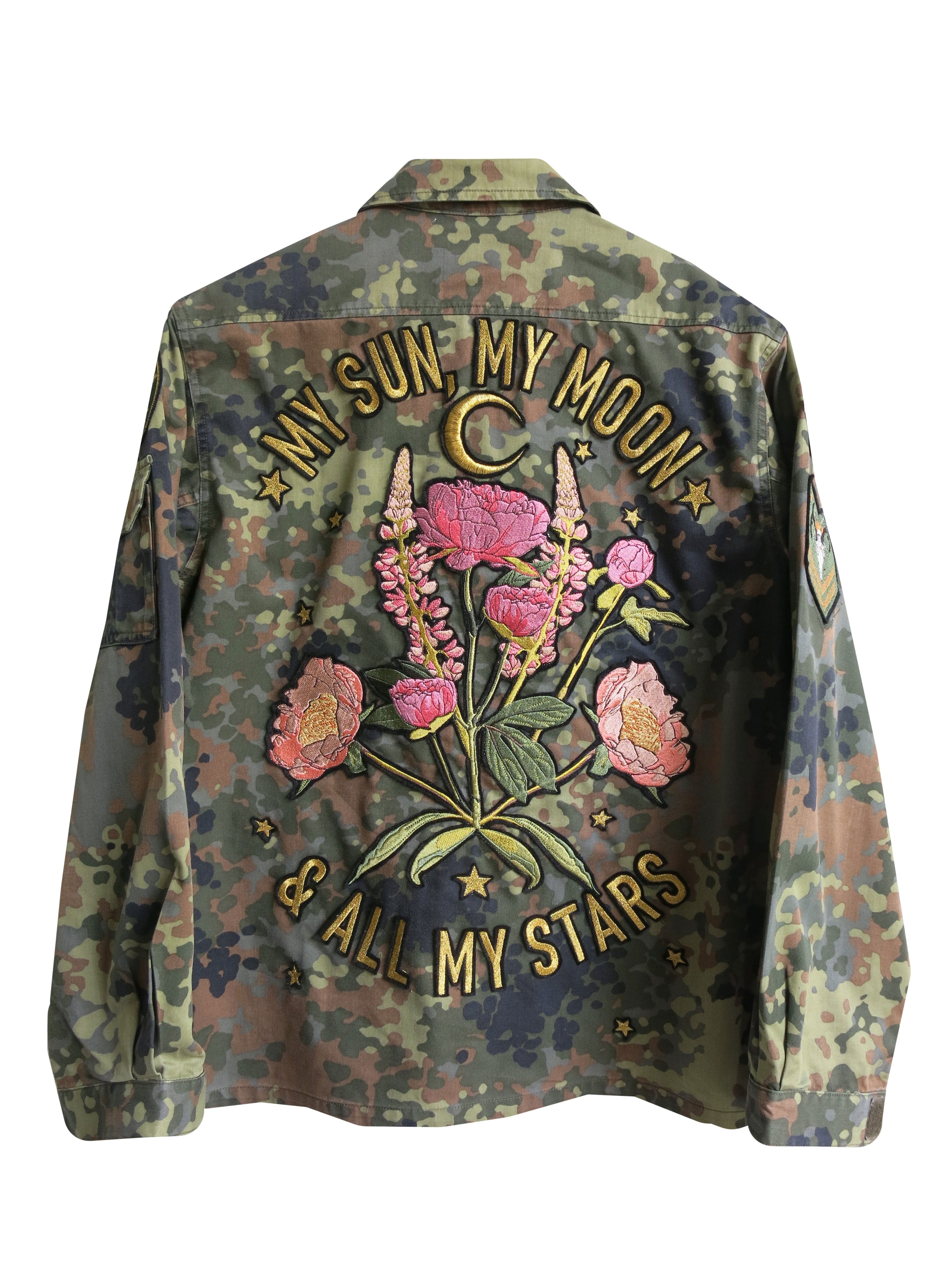 Back view of a camouflage military jacket with embroidered floral bouquet, crescent moon, and "MY SUN, MY MOON & ALL MY STARS" lettering, against a light wood-paneled wall.