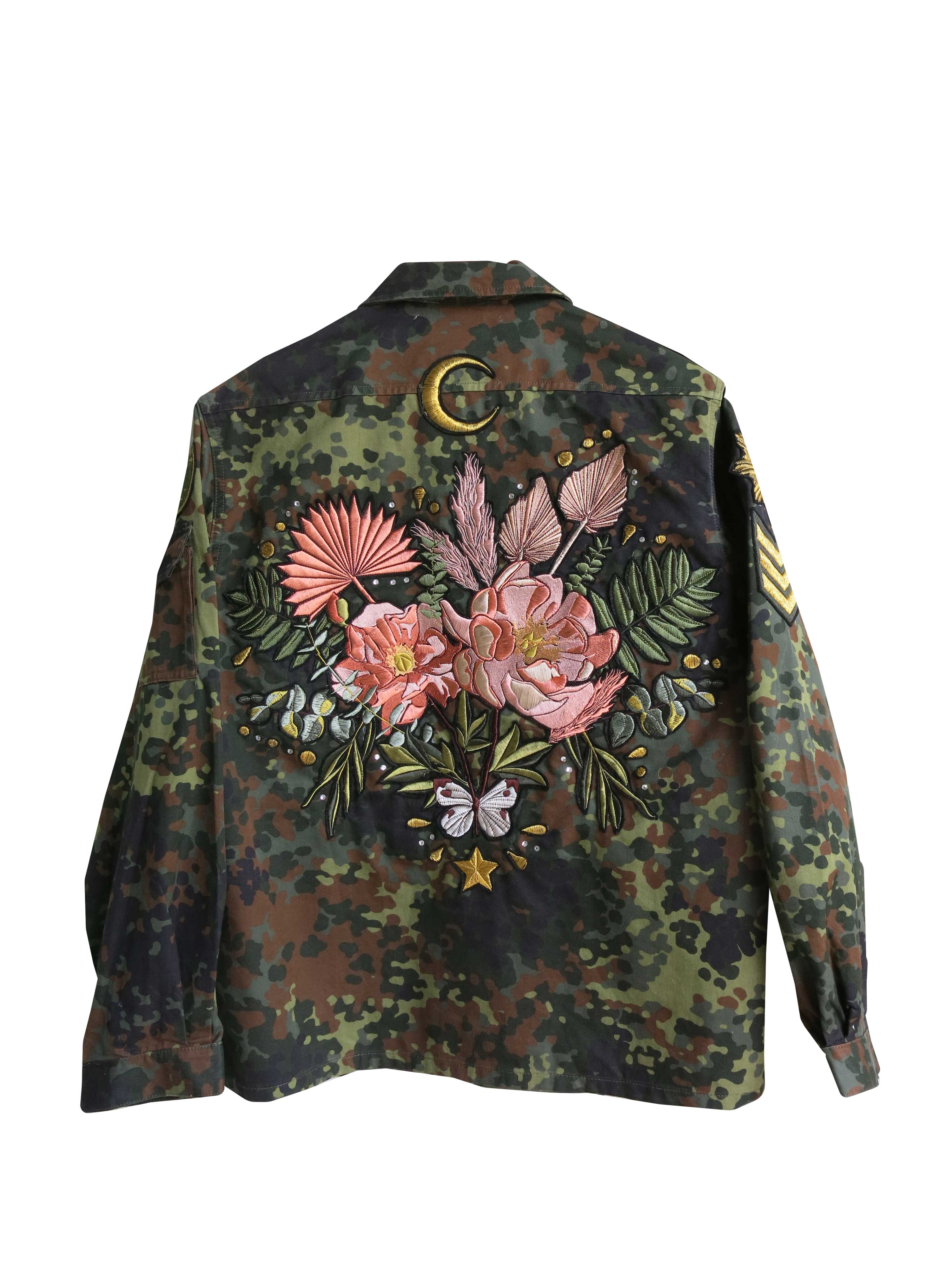 A camouflage military jacket with a large embroidered floral bouquet, crescent moon, butterfly, and botanical design on the back.