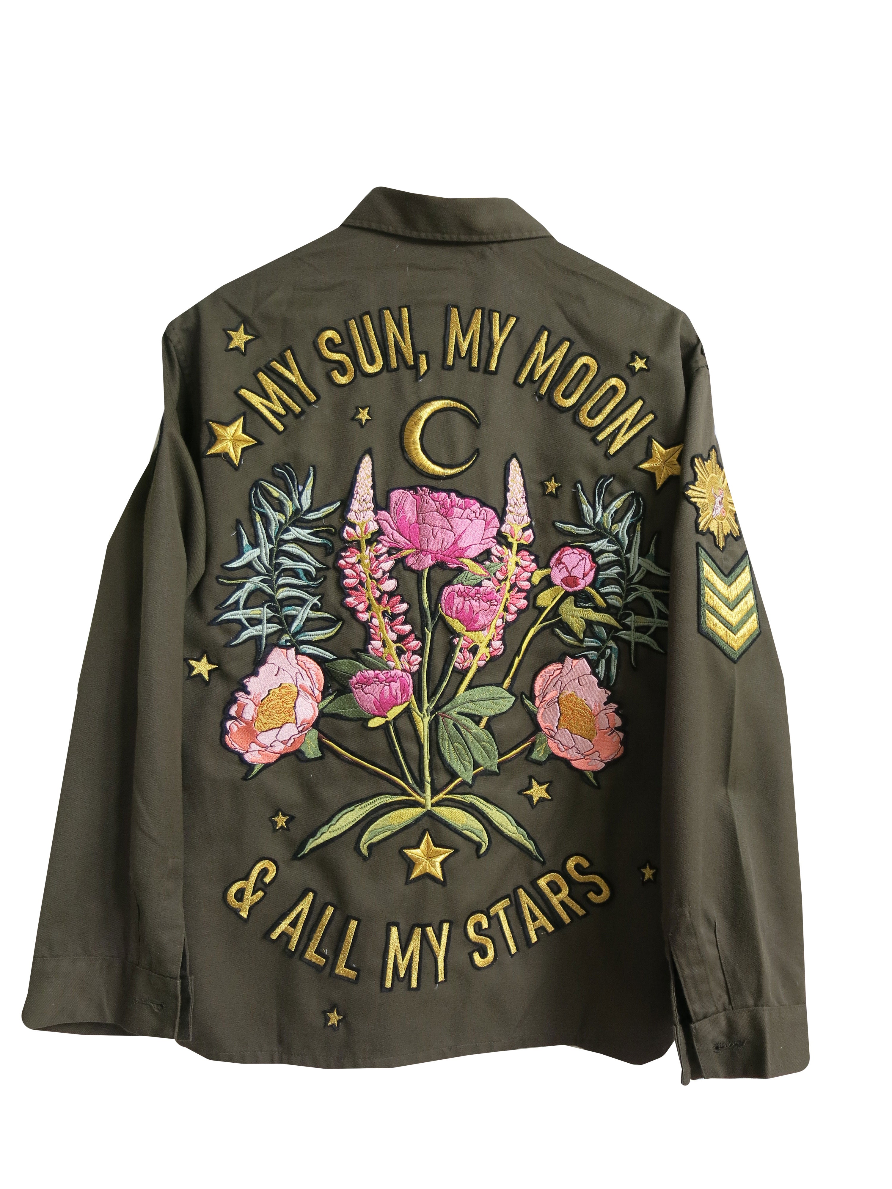Back view of an olive green military jacket with "MY SUN, MY MOON & ALL MY STARS" embroidered around a floral bouquet, crescent moon, and star design.