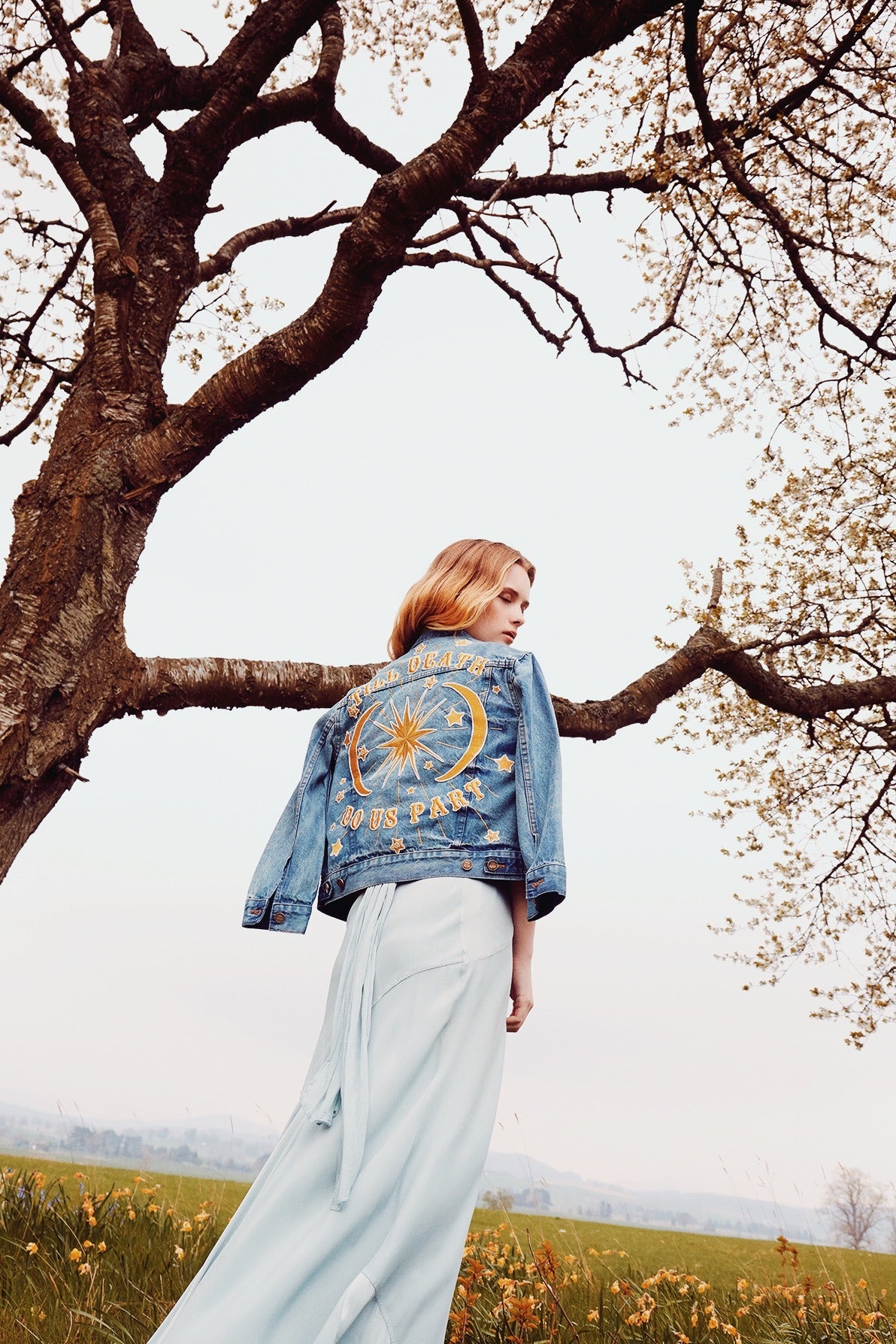 Woman wearing an embroidered denim jacket with a celestial "Till Death Do Us Part" design standing beneath a tree.