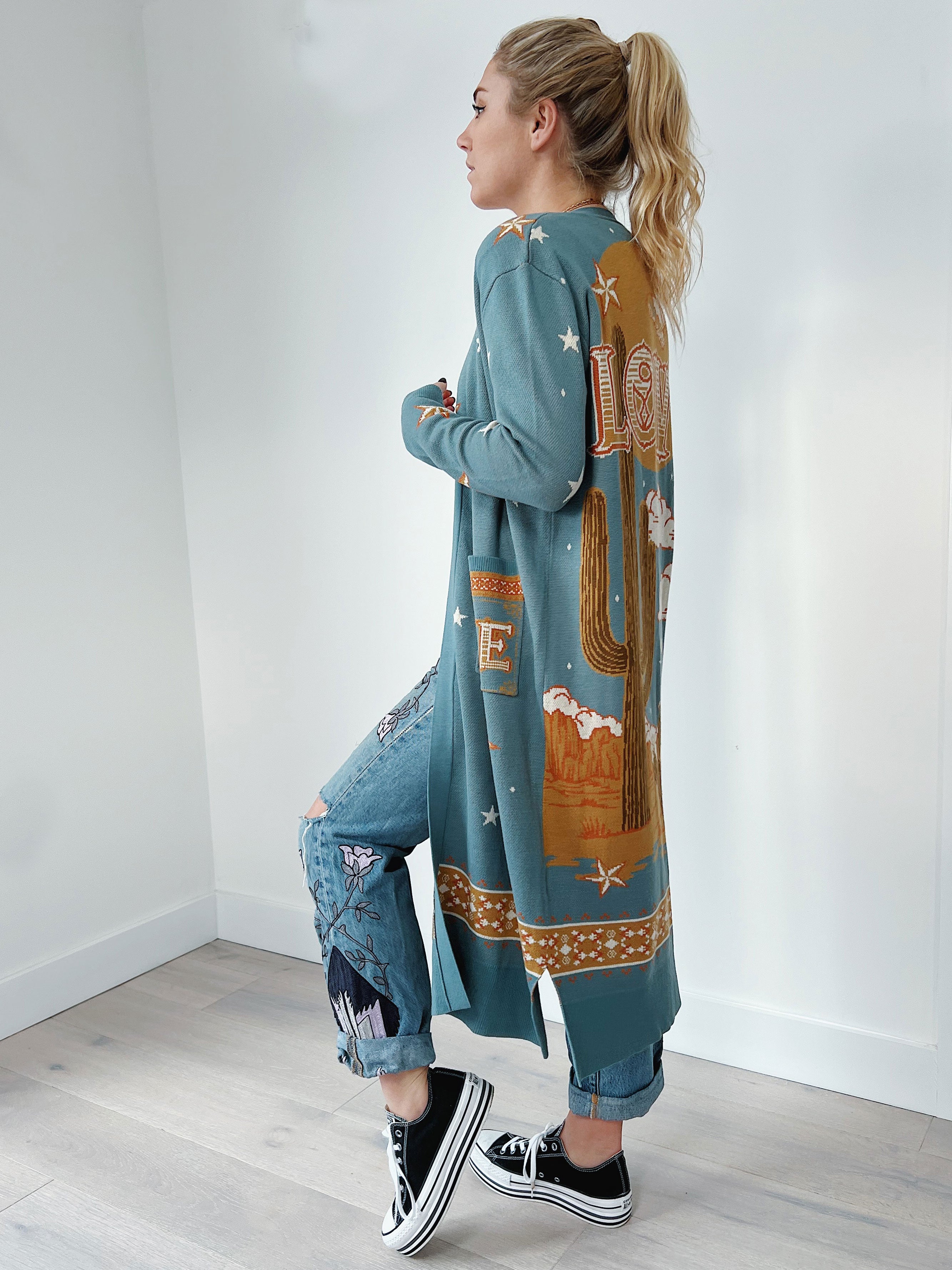 Side view of a person wearing a teal long knit cardigan featuring a western desert landscape knit design with a large cactus, stars, clouds, mesas, and the word "LOVE" on the back.