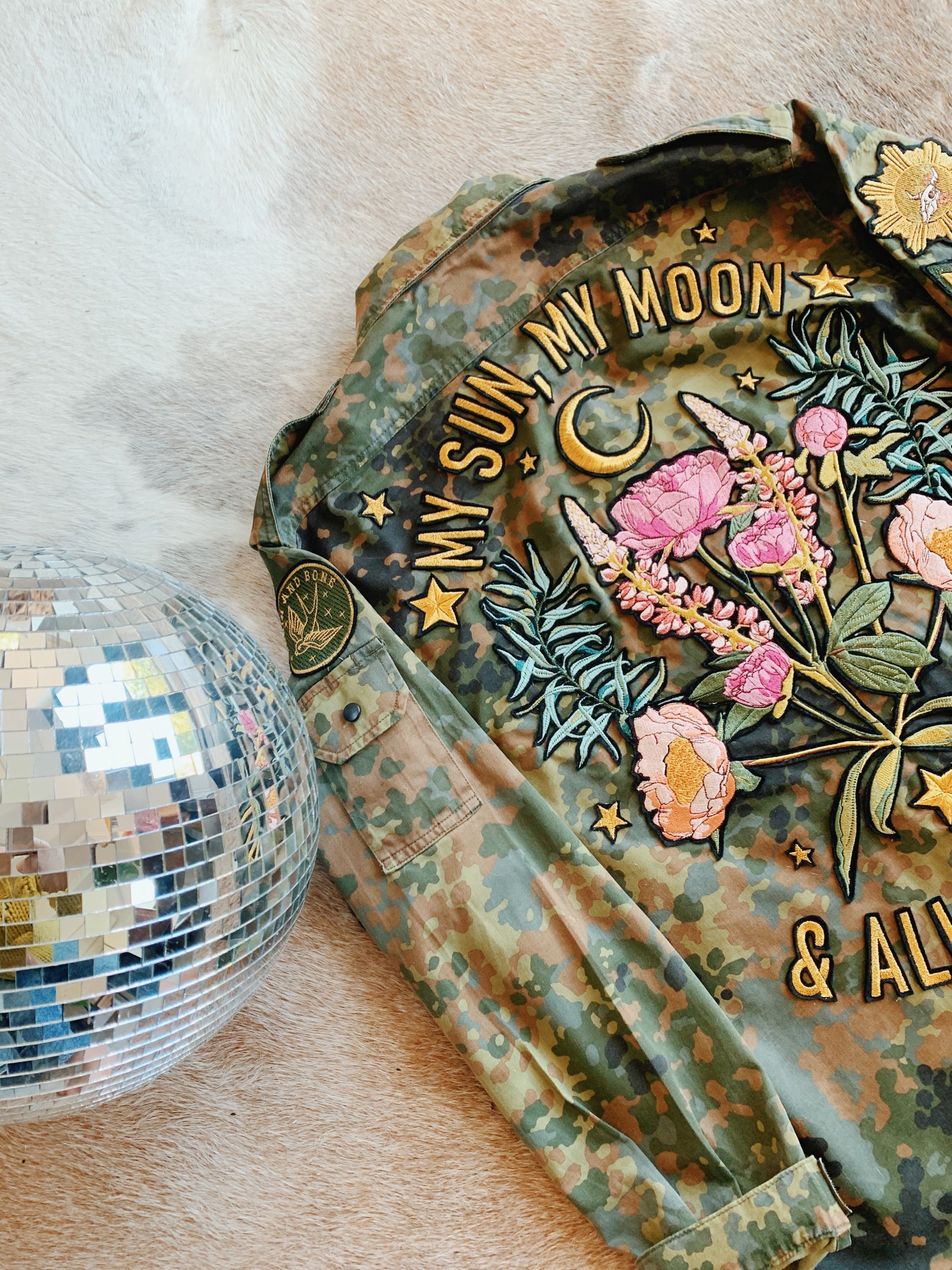 Flat lay of a camouflage military jacket with embroidered floral bouquet, crescent moon, and "MY SUN, MY MOON & ALL MY STARS" lettering, displayed beside a mirrored disco ball.