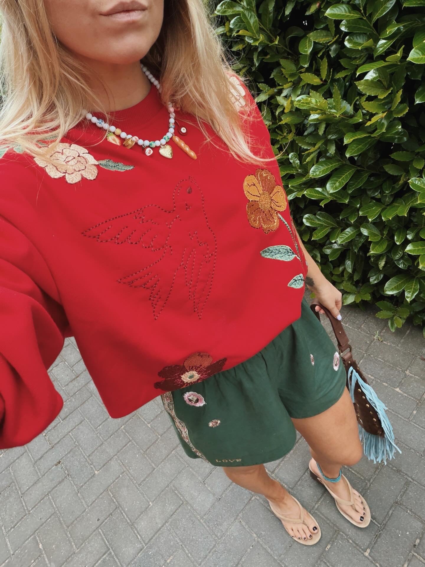Person wearing a red embroidered sweatshirt with floral and hummingbird embroidery, paired with green embroidered shorts.