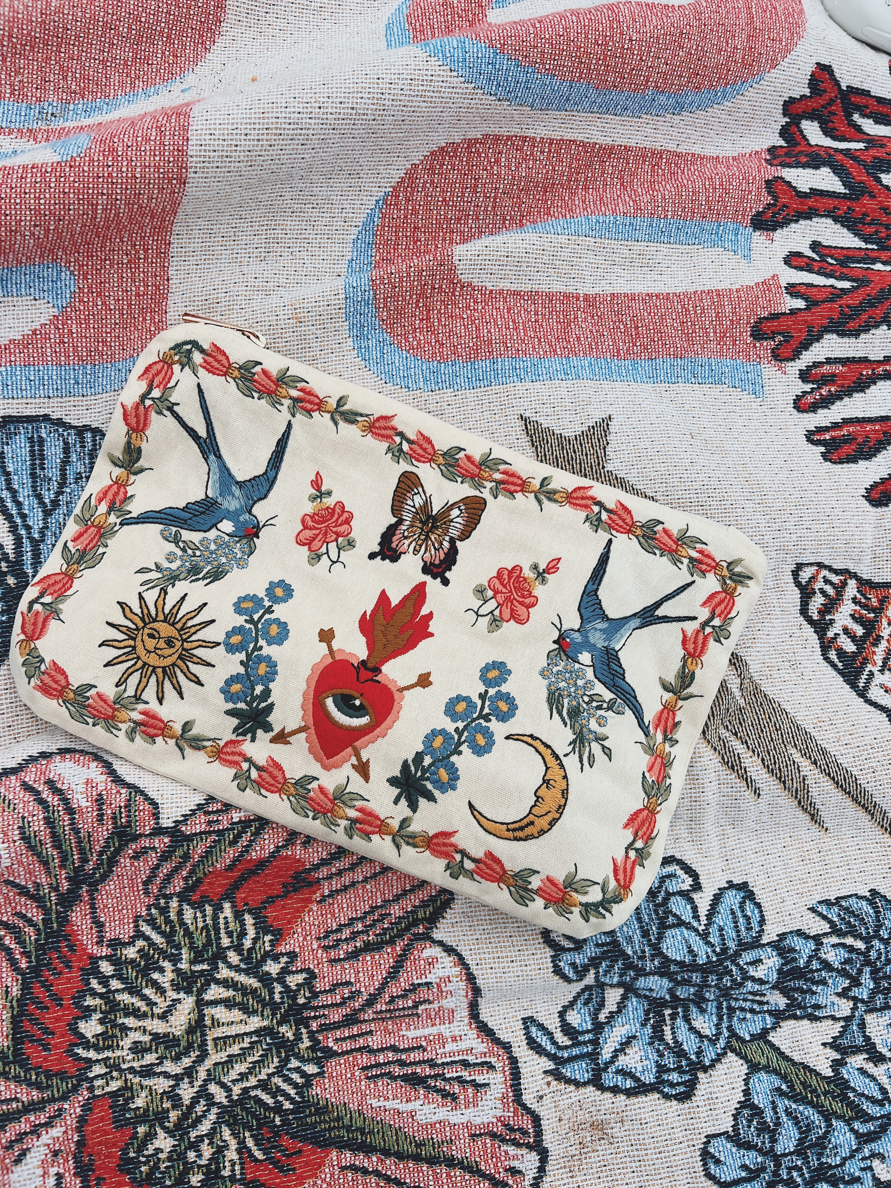 Cream zip pouch featuring embroidered swallows, florals, butterflies, a crescent moon, sun, and sacred heart motifs.