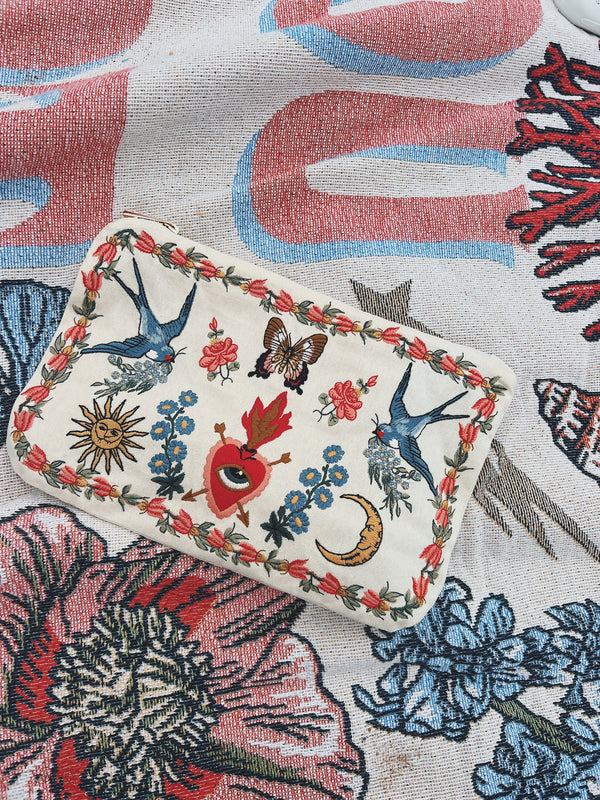 Cream zip pouch featuring embroidered swallows, florals, butterflies, a crescent moon, sun, and sacred heart motifs.