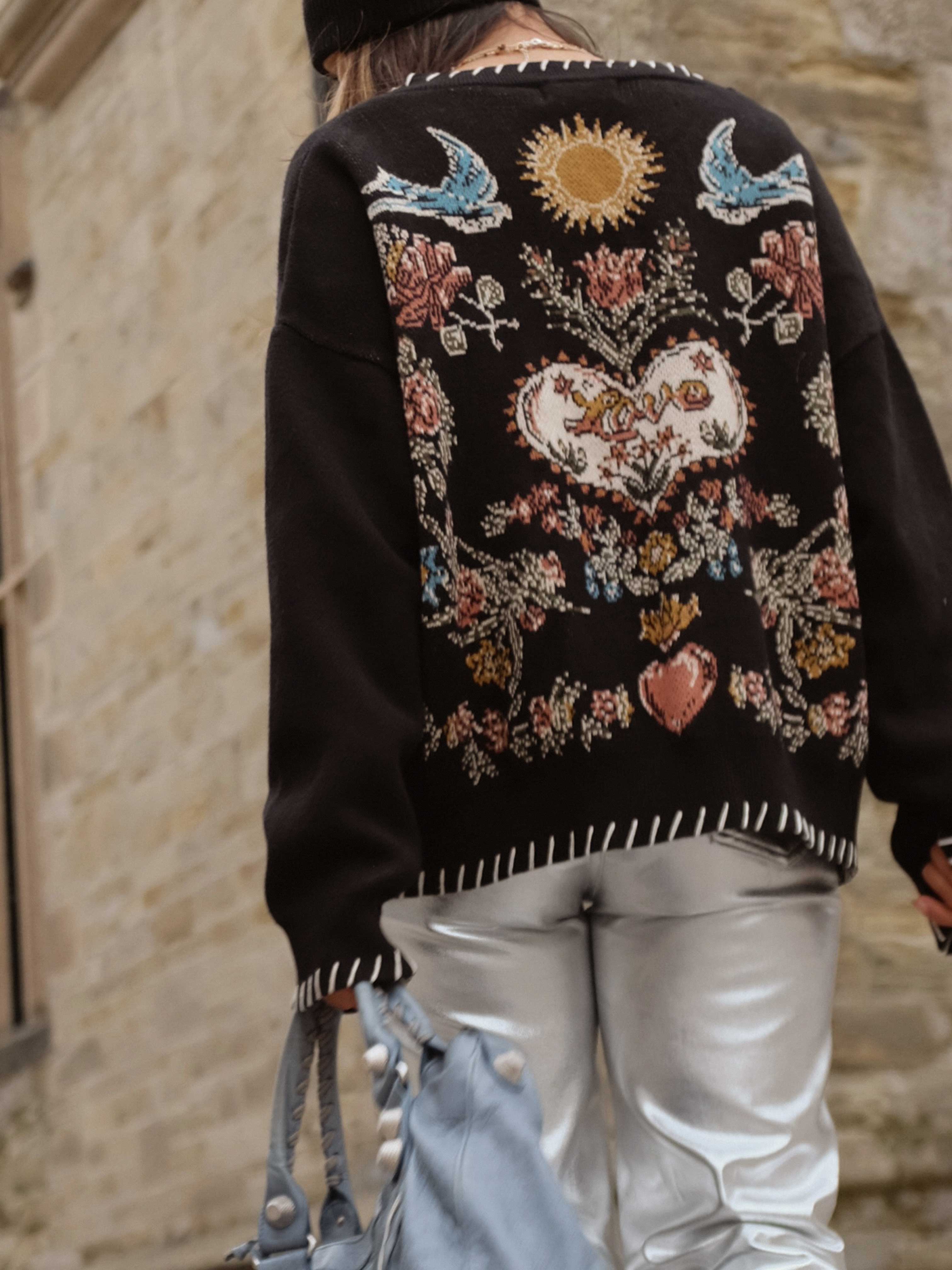 Back view of a person wearing a black knit cardigan featuring a heart, flowers, birds, and sun knit design.