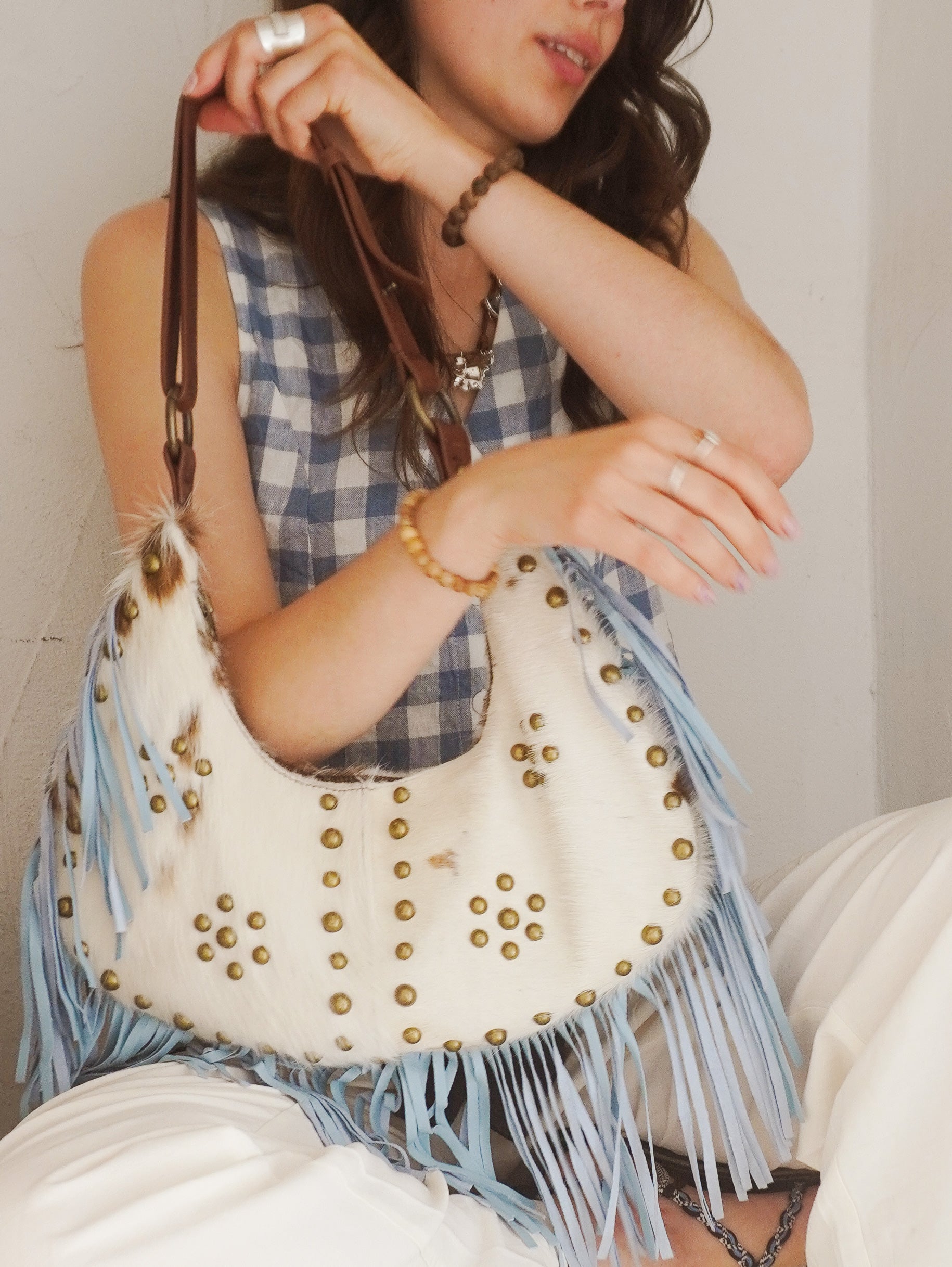 Person carrying a cream and brown cowhide leather shoulder bag with light blue suede fringe, brass stud detailing, and a brown leather strap.