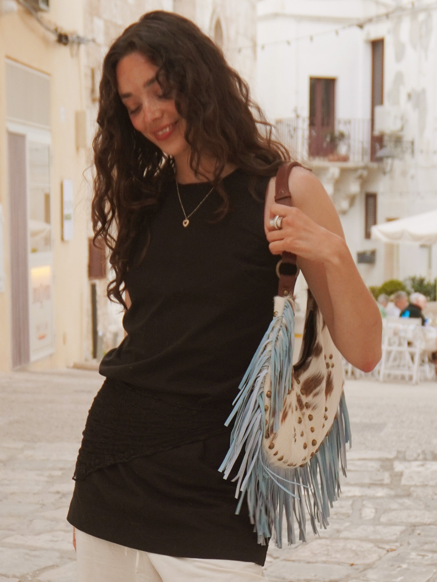Person carrying a cream and brown cowhide leather shoulder bag with light blue suede fringe, brass stud detailing, and a brown leather strap.