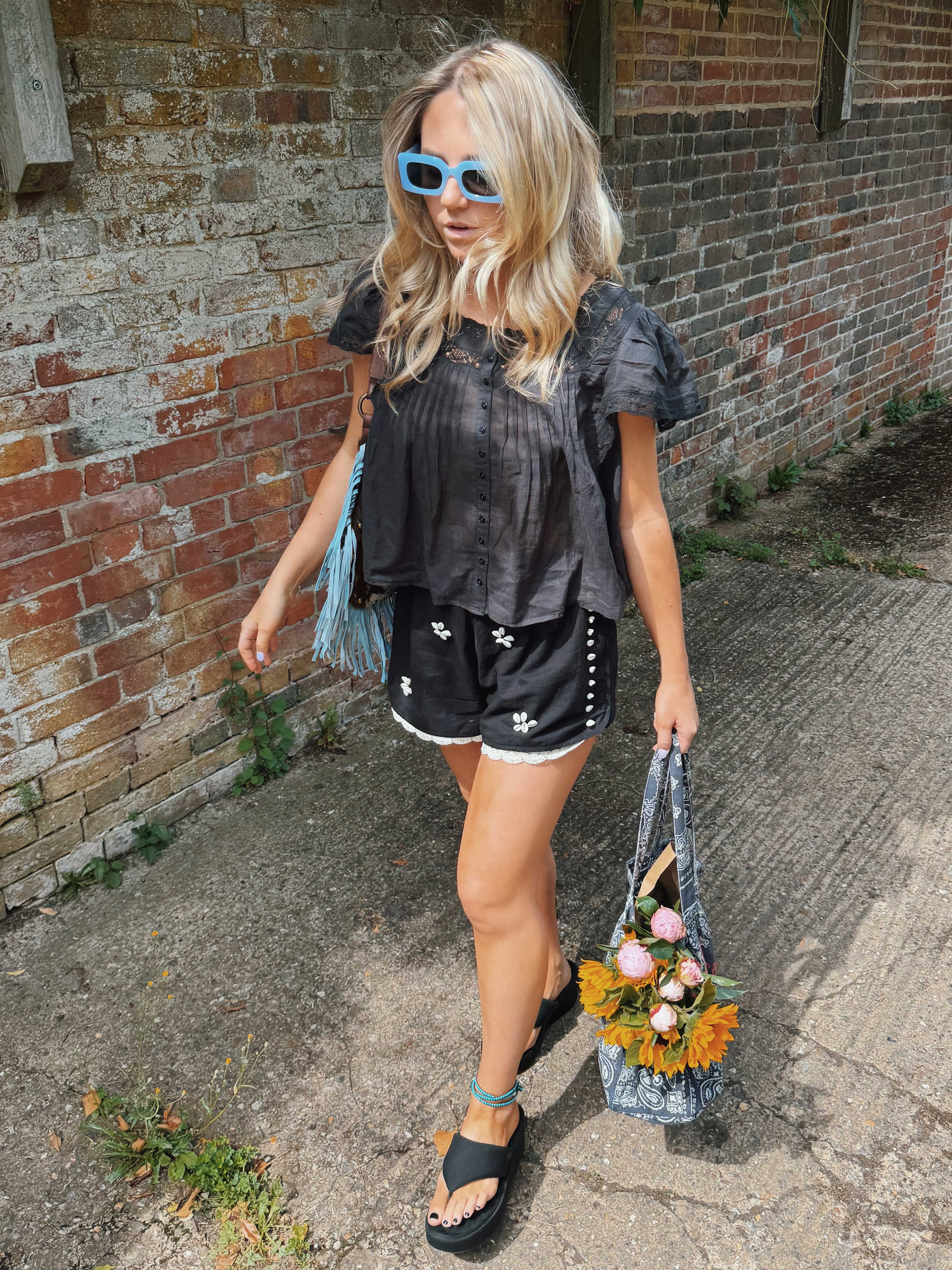 Woman wearing black embroidered shorts with white floral embroidery, shell side detailing, and lace trim, paired with a black flutter sleeve blouse and carrying a floral tote bag.