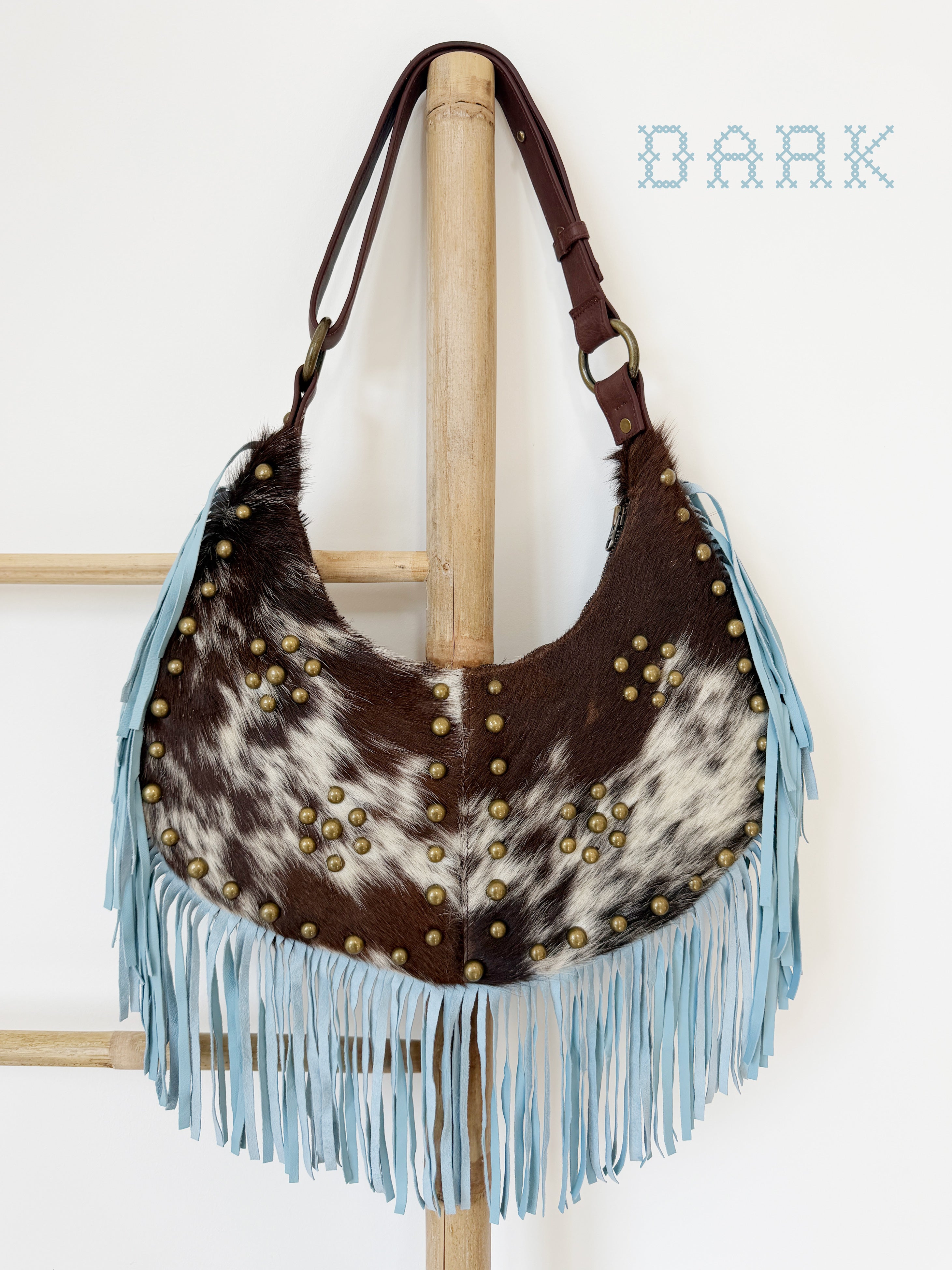 Dark brown and white cowhide leather shoulder bag with light blue suede fringe, brass stud detailing, and a brown leather strap.