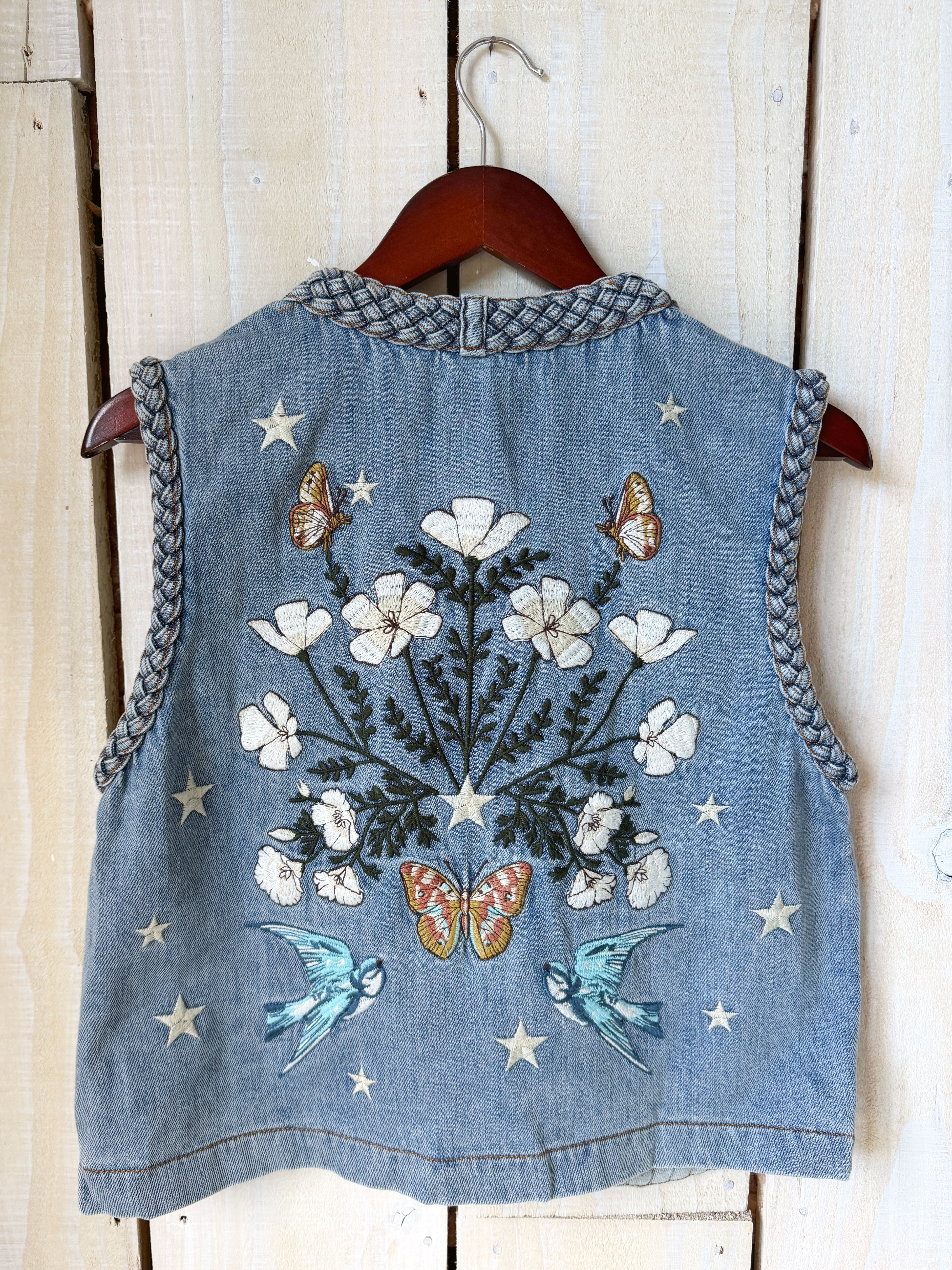 Back view of a blue denim vest with white floral embroidery, butterflies, birds, and star details.