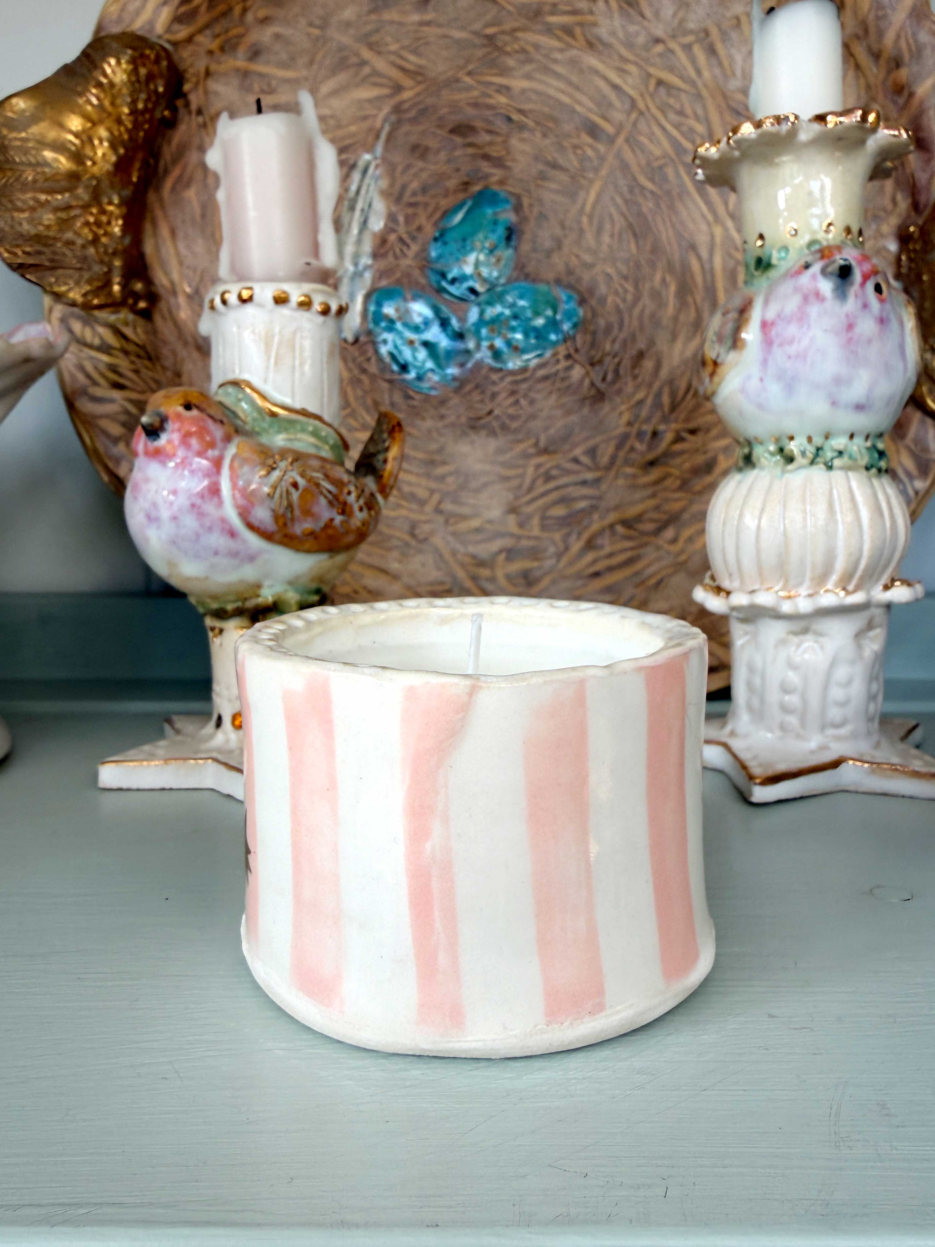 A handmade ceramic candle holder with vertical pink and white stripes sits on a light blue-gray surface, styled with other decorative ceramic bird and birdhouse items in the background.