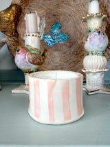 A handmade ceramic candle holder with vertical pink and white stripes sits on a light blue-gray surface, styled with other decorative ceramic bird and birdhouse items in the background.