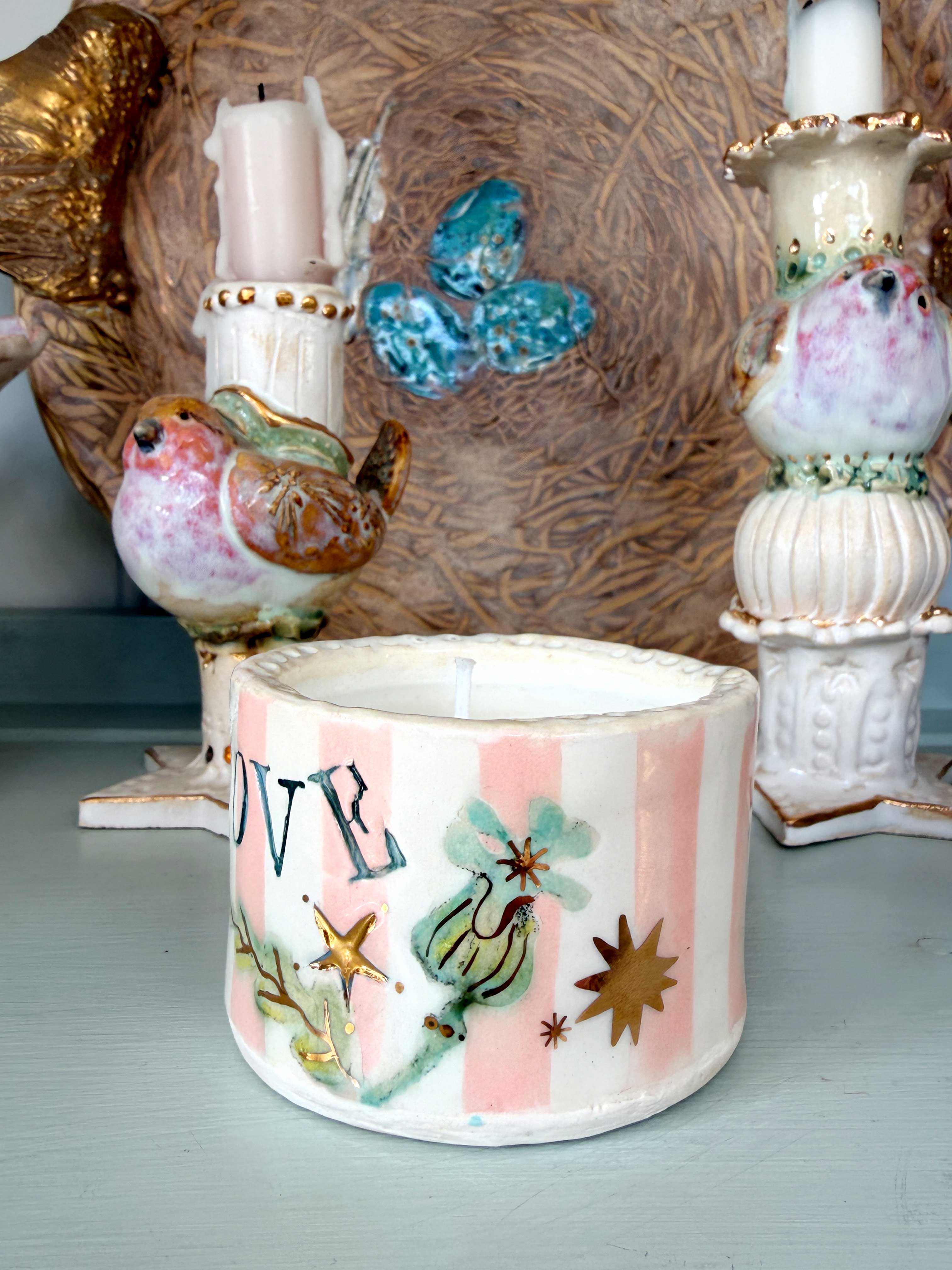 A handmade ceramic candle holder with pink and white stripes, decorated with green birds, gold stars, and the word 'LOVE', shown in a styled tabletop setting with other decorative ceramic items.