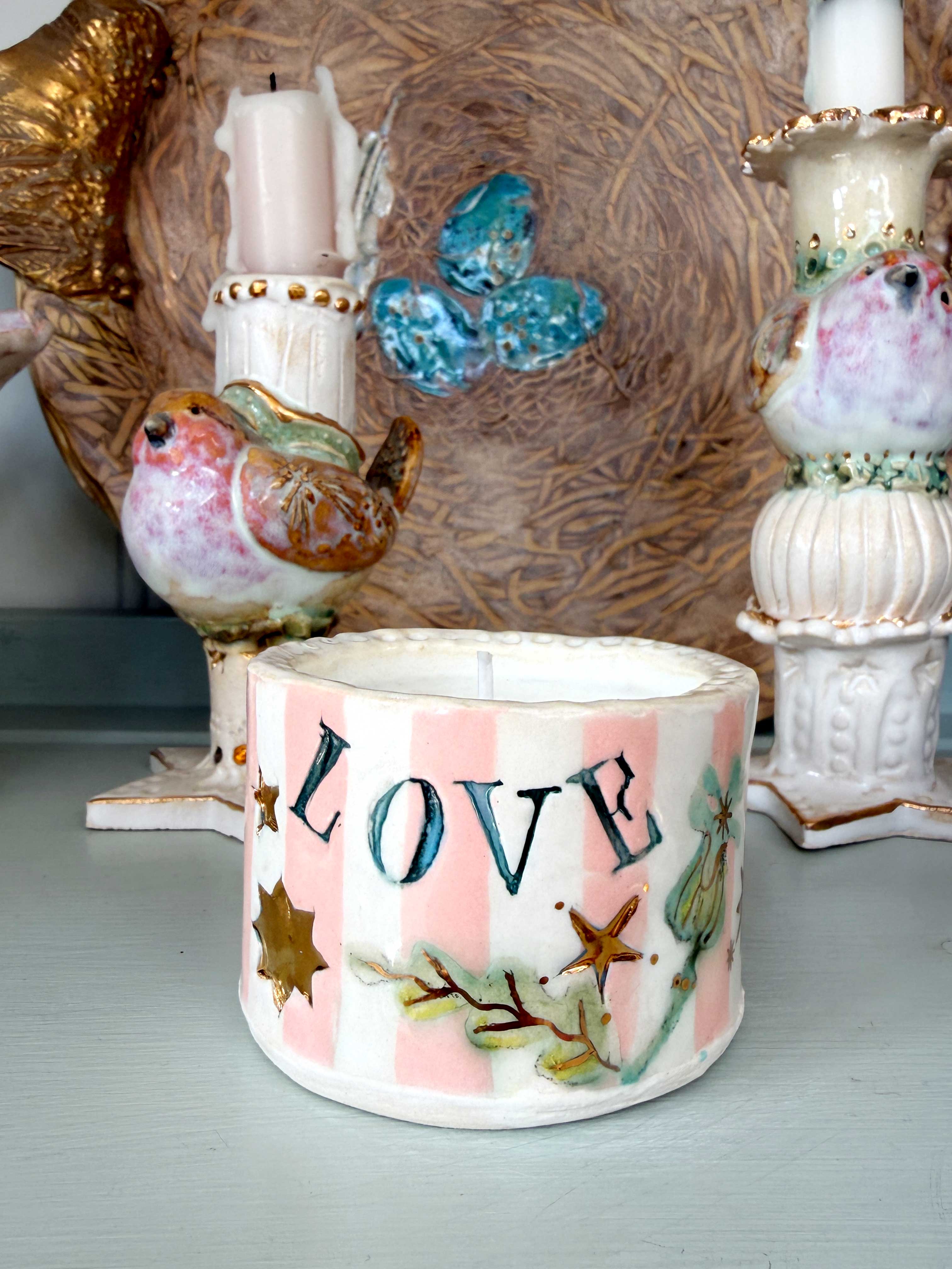 A handmade ceramic candle holder with pink and white vertical stripes, decorated with the word 'LOVE' in blue letters, gold stars, and green leaves. It is styled on a tabletop with other decorative ceramic items in the background.