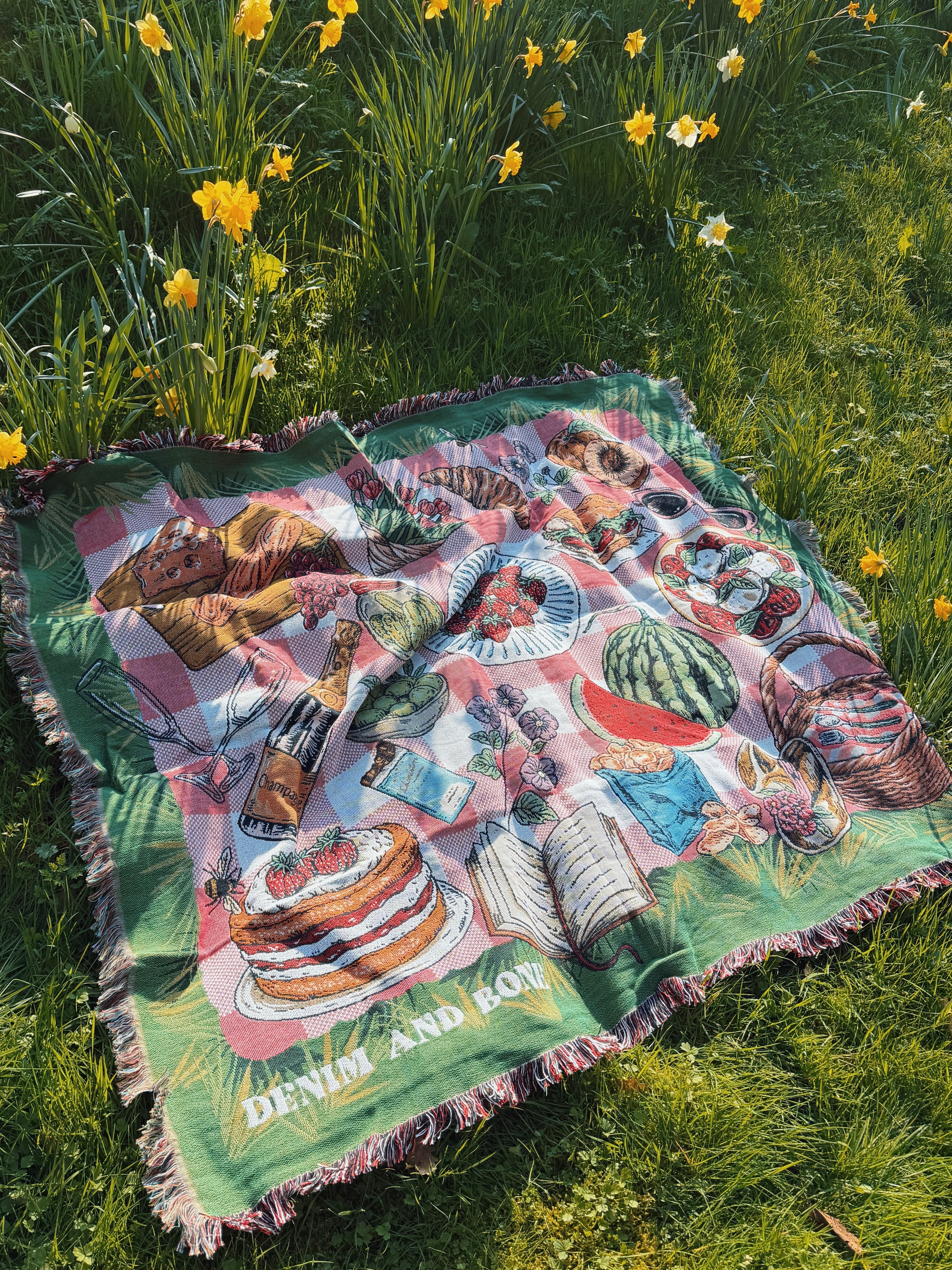 Woven picnic blanket with a pink gingham picnic design laid on grass beside blooming daffodils.