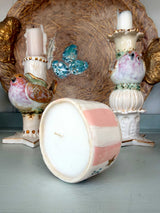 A handmade ceramic candle shown on its side in the foreground, featuring pink and white stripes with a small brown bird detail. In the background, two matching bird-shaped candlesticks and a decorative woven basket are visible.