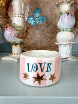 A handmade ceramic candle holder with the word 'LOVE' in blue letters, decorated with gold star cutouts and pink and white stripes, displayed on a shelf between other decorative bird figurines.