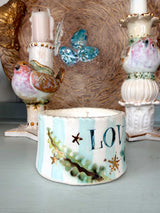 A decorative ceramic candle holder or candle container in the foreground, painted with light blue and white vertical stripes, green pine branches, gold stars, and the partial word "LOVE" in dark blue. Two ornate white bird candlesticks and a rustic woven backdrop are visible behind it.