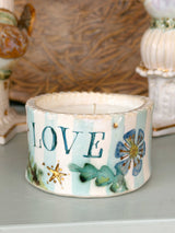 A small ceramic candle holder or candle container with the word “LOVE” painted in teal, decorated with blue flowers, green leaves, and gold stars on a white and light blue striped background, set on a tabletop with a softly blurred background.