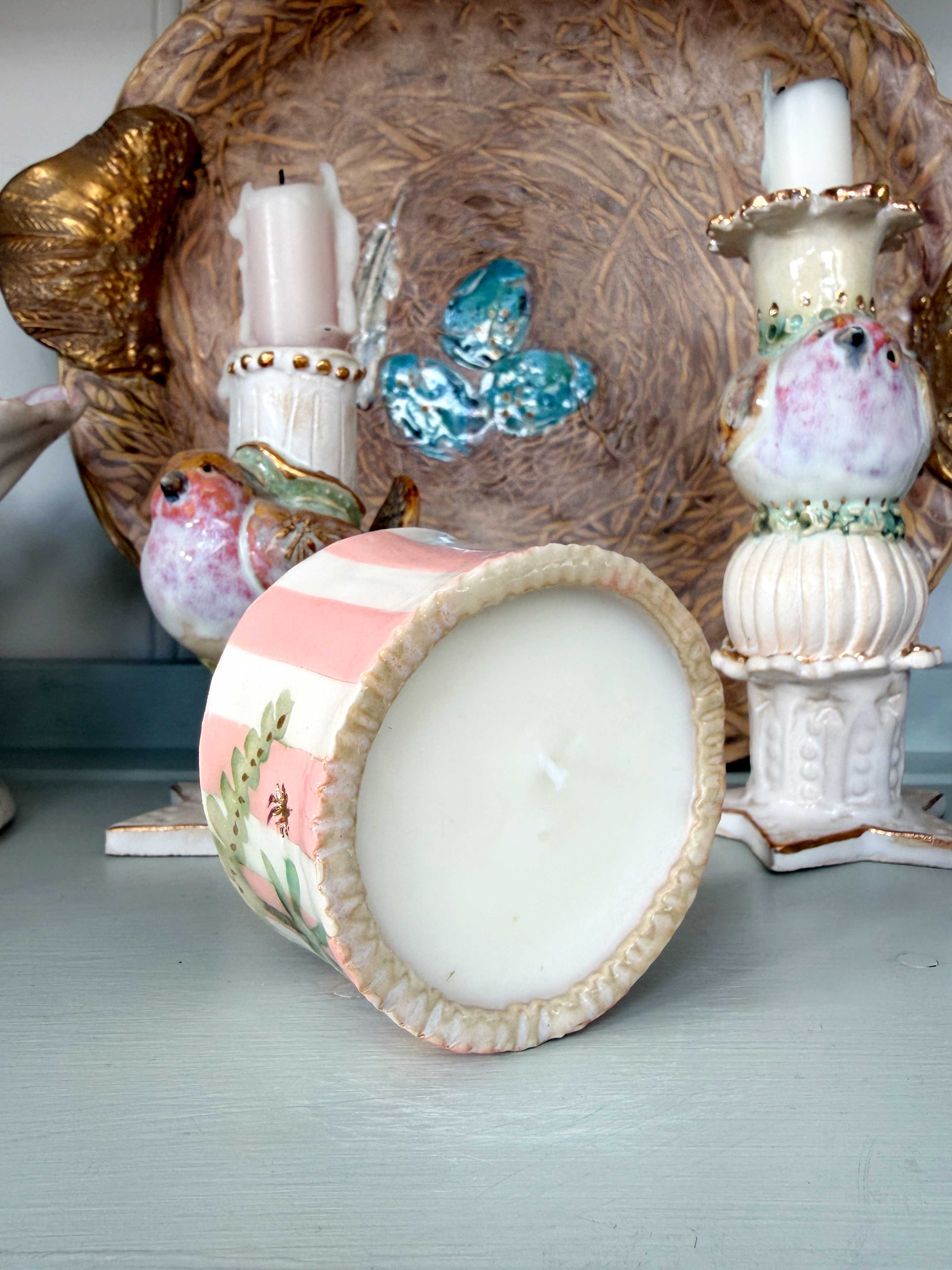 A pastel pink and white striped ceramic candle holder with a floral design, shown tipped over to display the white wax inside. The background features other decorative vintage-style ceramic objects on a light blue-gray surface.