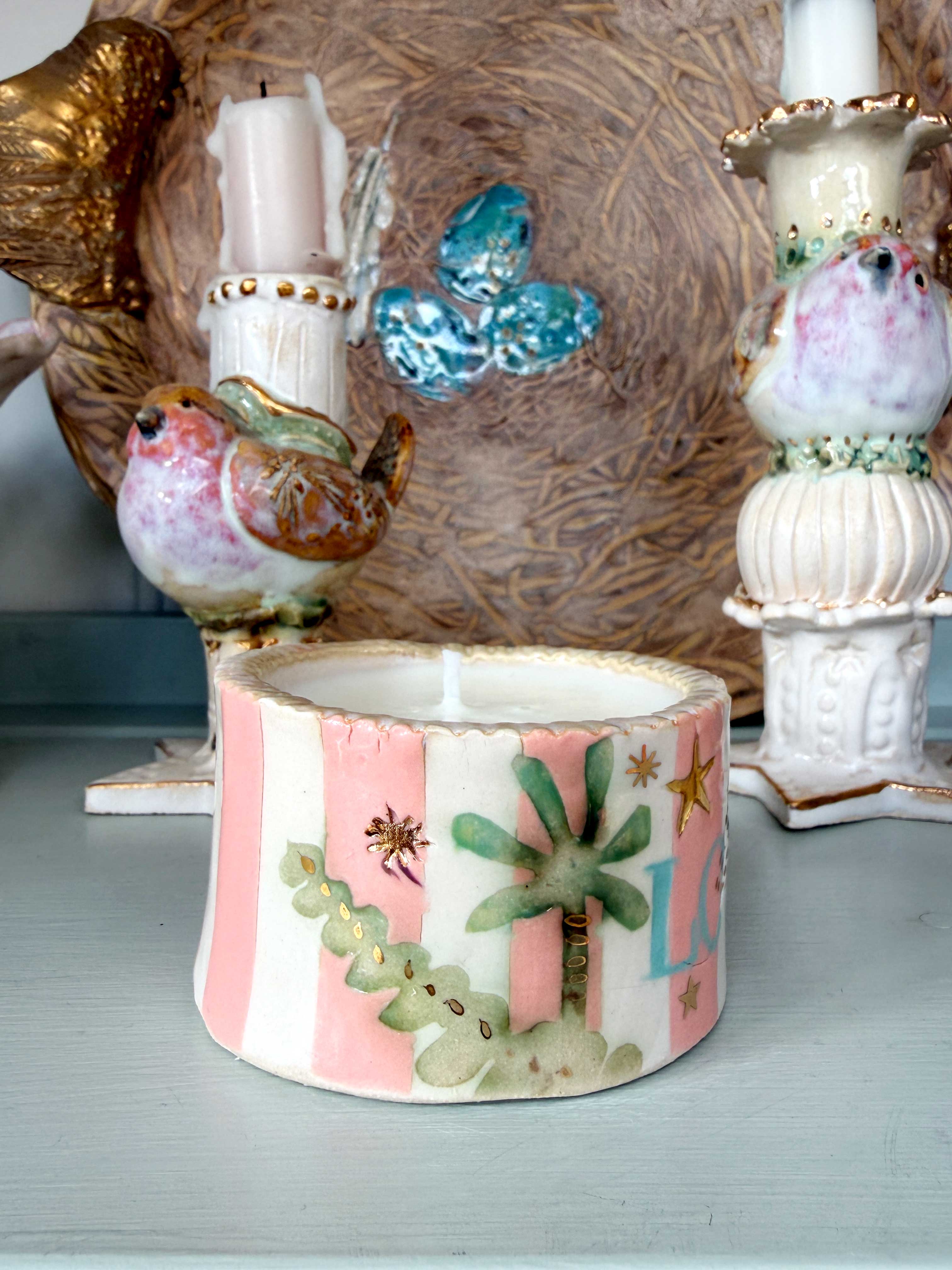 A handmade ceramic candle holder featuring pink and white vertical stripes with painted green palm trees, cactus, and star details, displayed on a light gray tabletop.