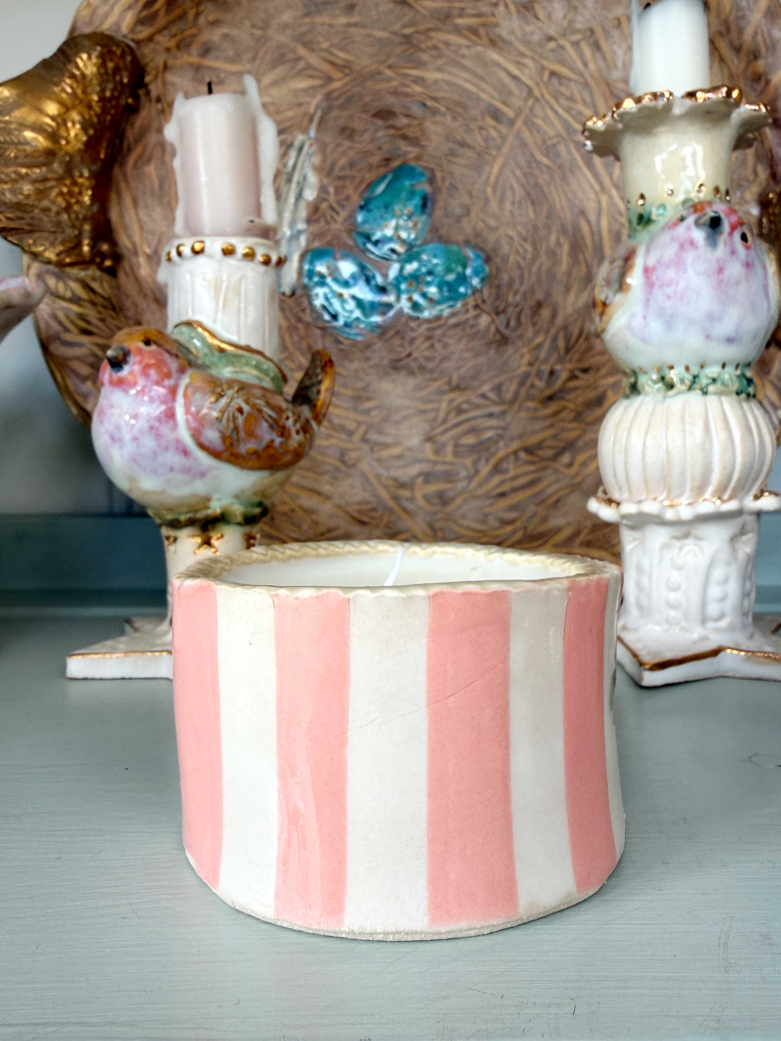 A pink and white vertically striped ceramic candle holder with a bird-shaped candle holder and a blue butterfly decoration in the background.