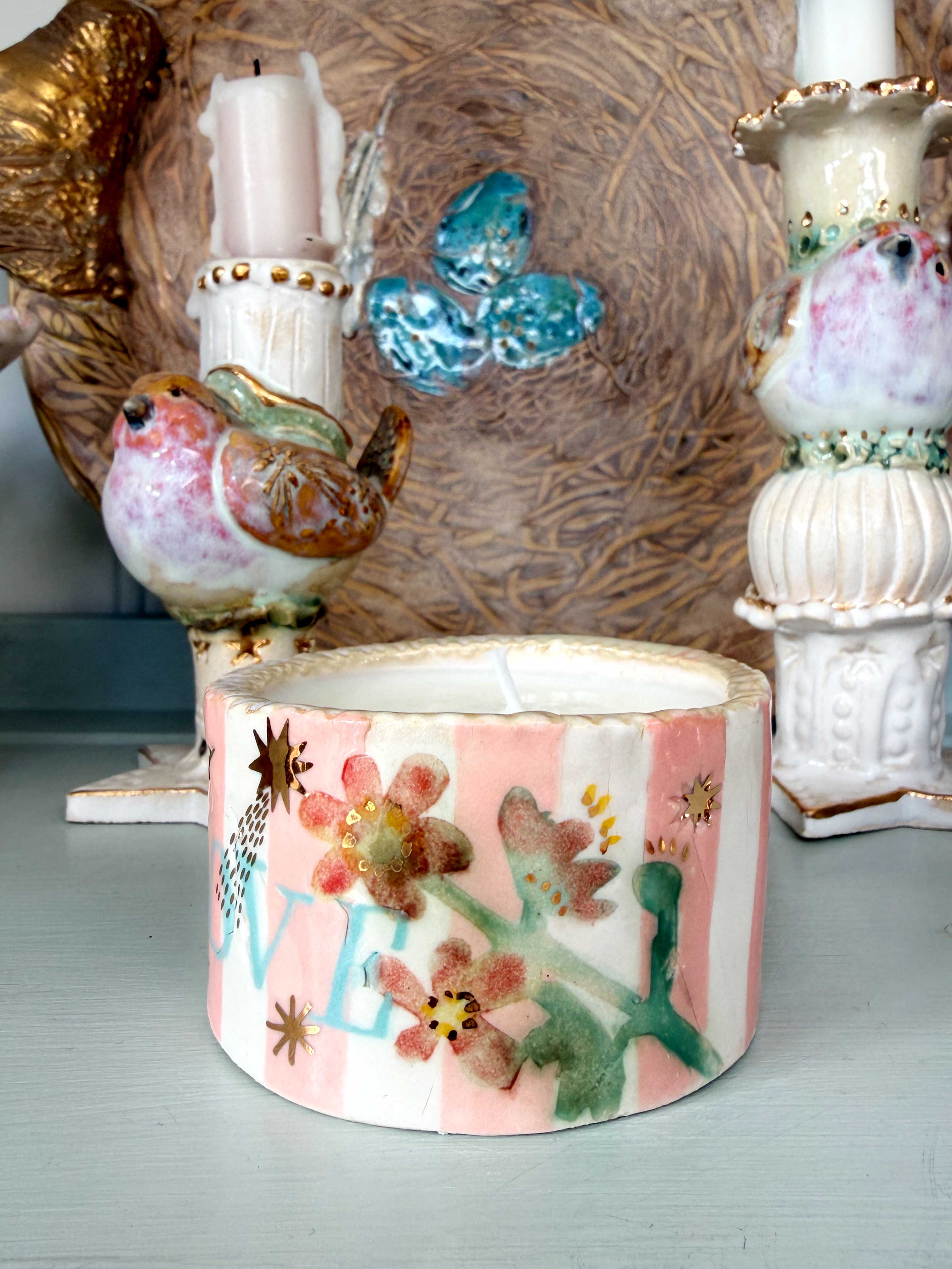 A handmade ceramic candle holder shaped like a bird, decorated with pink and white stripes, colorful flowers, and green leaves, shown on a tabletop with other decorative items in the background.