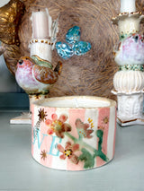 A handmade ceramic candle holder shaped like a bird, decorated with pink and white stripes, colorful flowers, and green leaves, shown on a tabletop with other decorative items in the background.