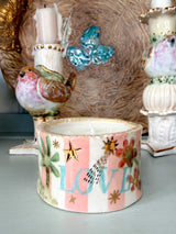 A small decorative ceramic candle holder with a pink and white striped pattern, gold stars, a bird design, and the word 'LOVE' in light blue, shown in a styled tabletop setting with other ceramic decor.