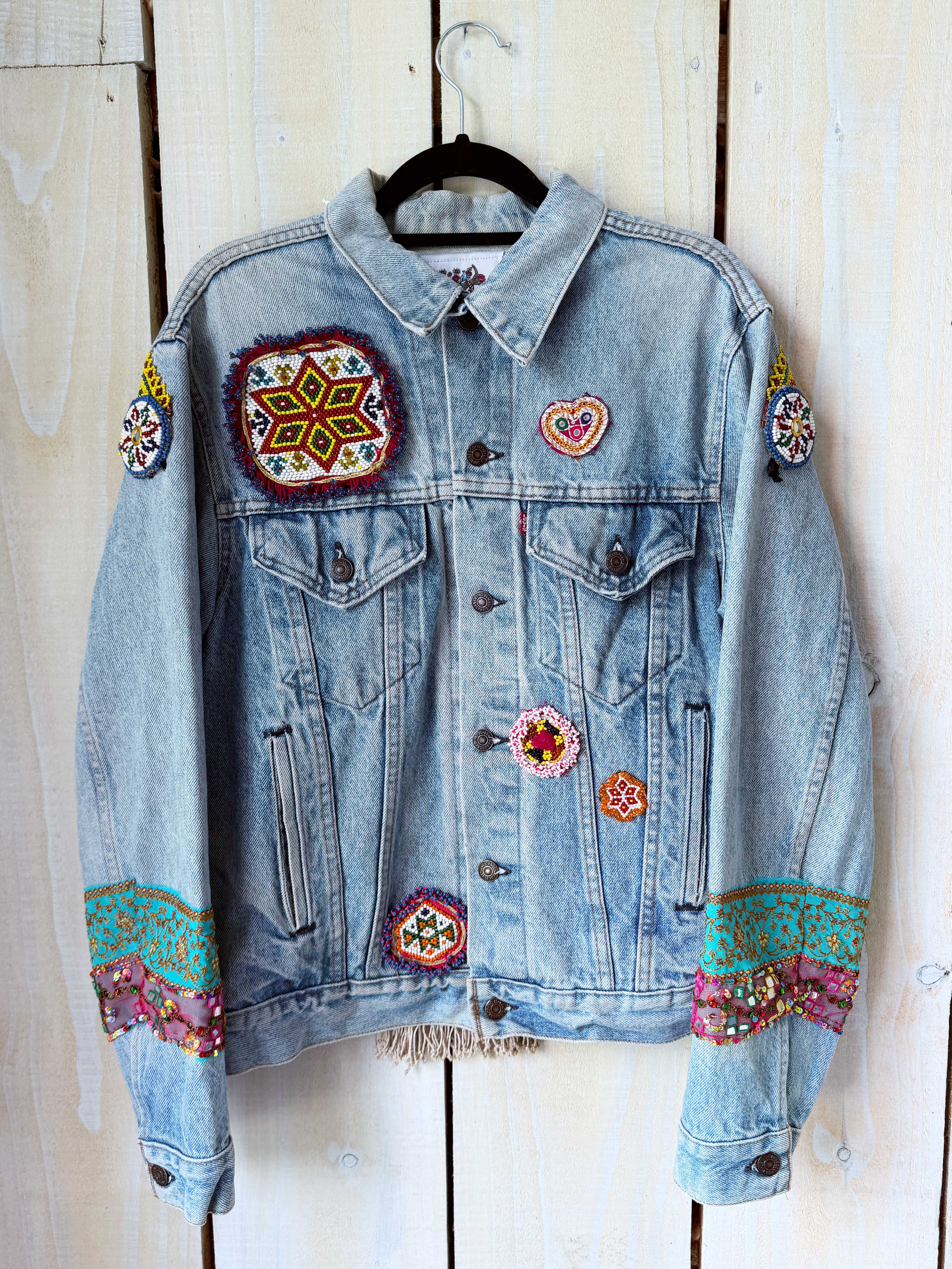 Light blue denim jacket with colourful embroidered and beaded patches, shown from the front on a white rustic wooden wall