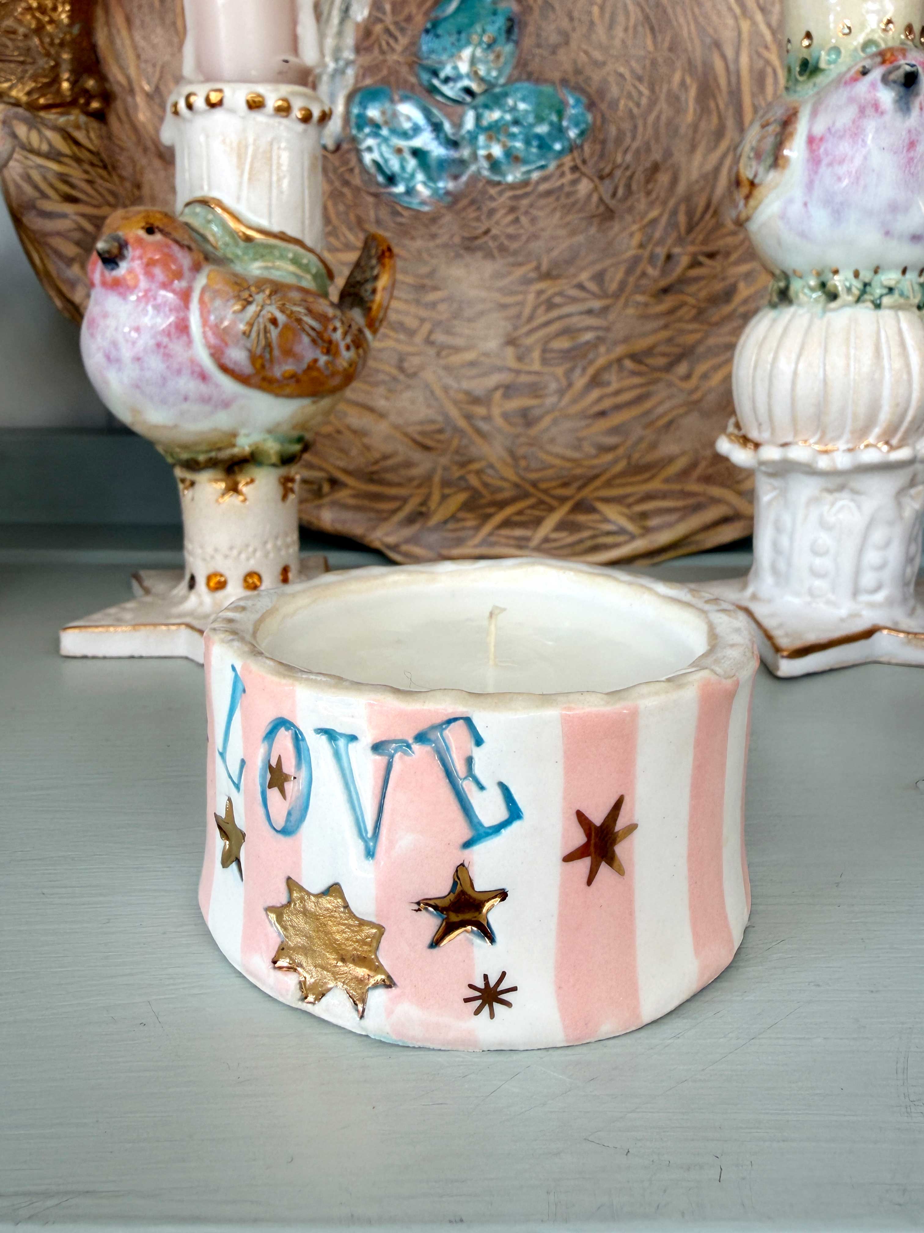 A small ceramic candle holder or candle base with pink and white vertical stripes, the word "LOVE" in blue letters, and gold star and leaf motifs, shown in front of decorative bird and bird figurines.