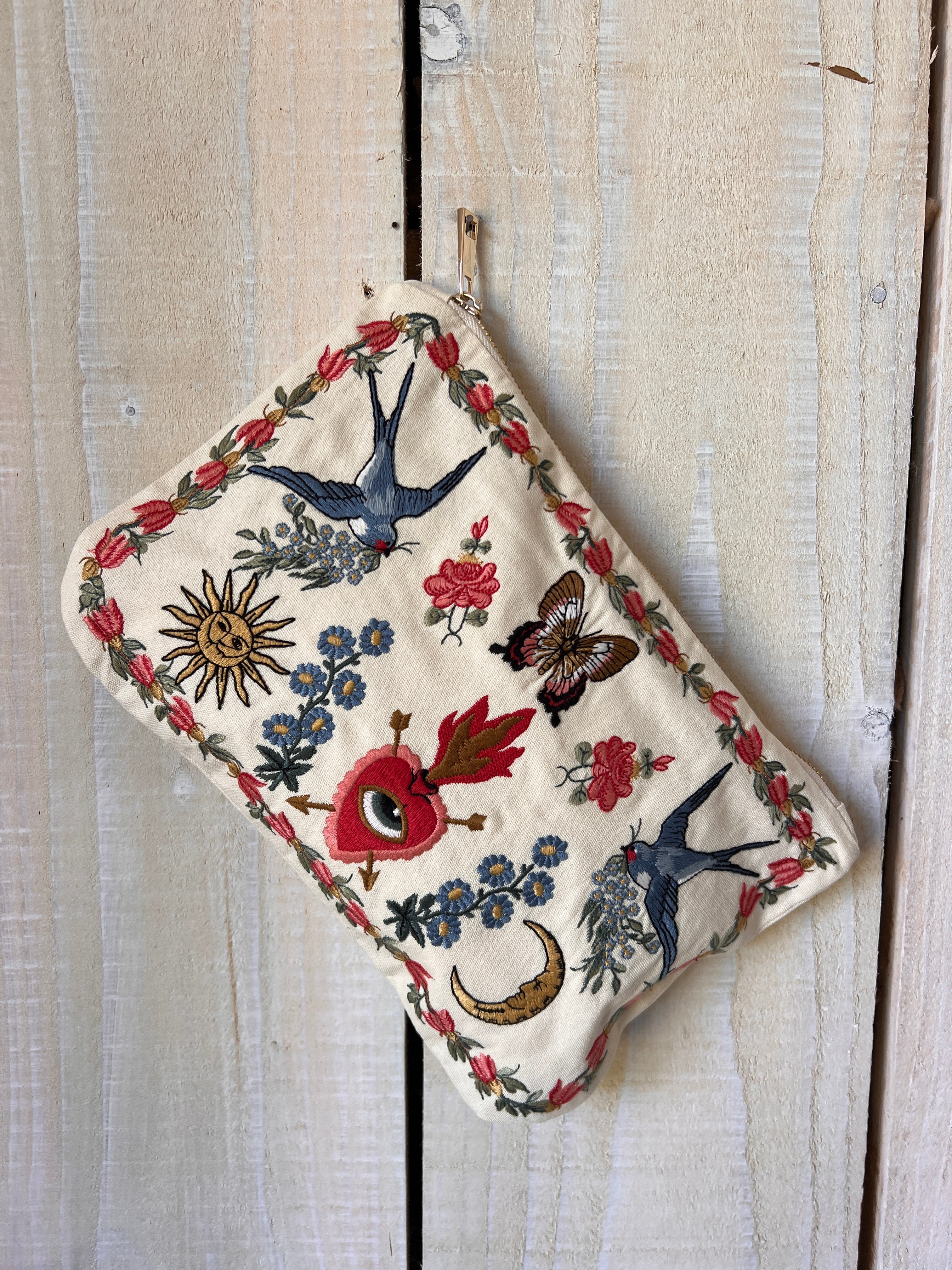 Cream zip pouch featuring embroidered swallows, florals, butterflies, a crescent moon, sun, and sacred heart motifs.