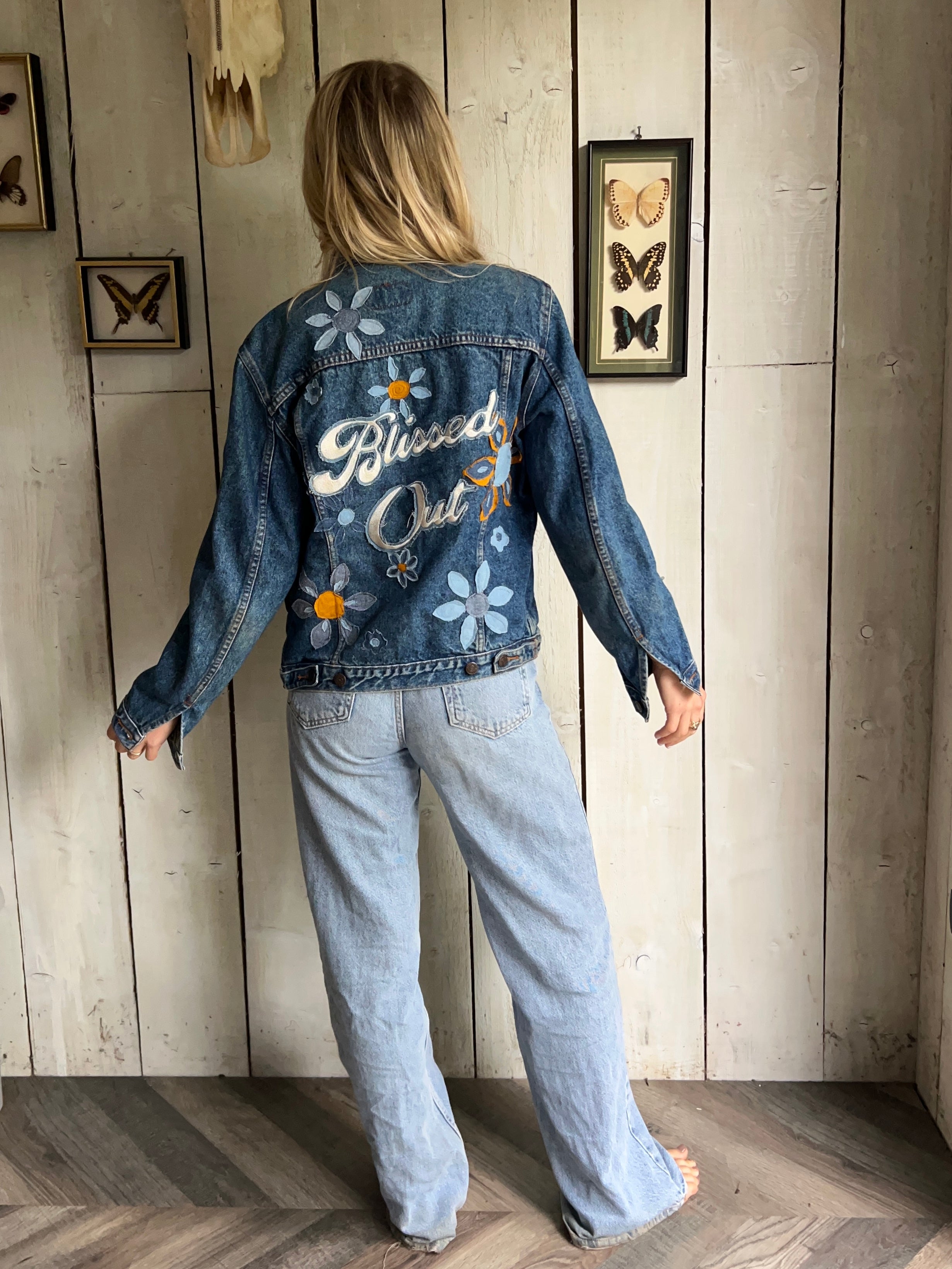 Blue denim jacket with "Blissed Out" embroidered back design and floral embroidery, shown from the back.