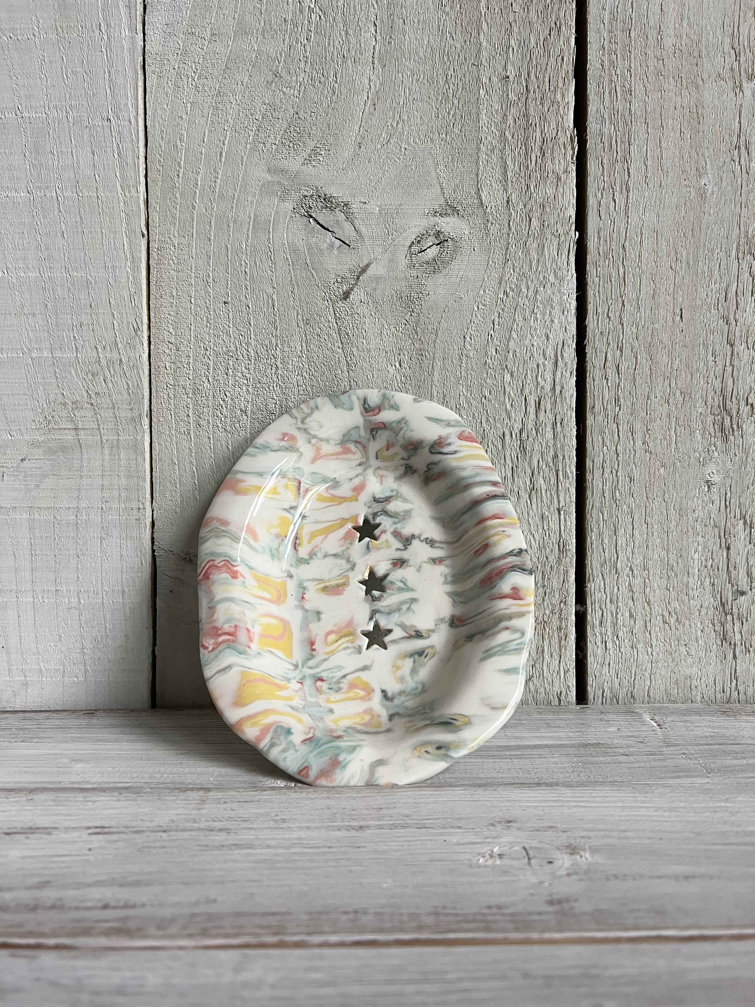 Handmade oval ceramic trinket dish with a pastel marbled glaze, scalloped rim, and three star-shaped cut-outs displayed on a rustic wooden shelf.