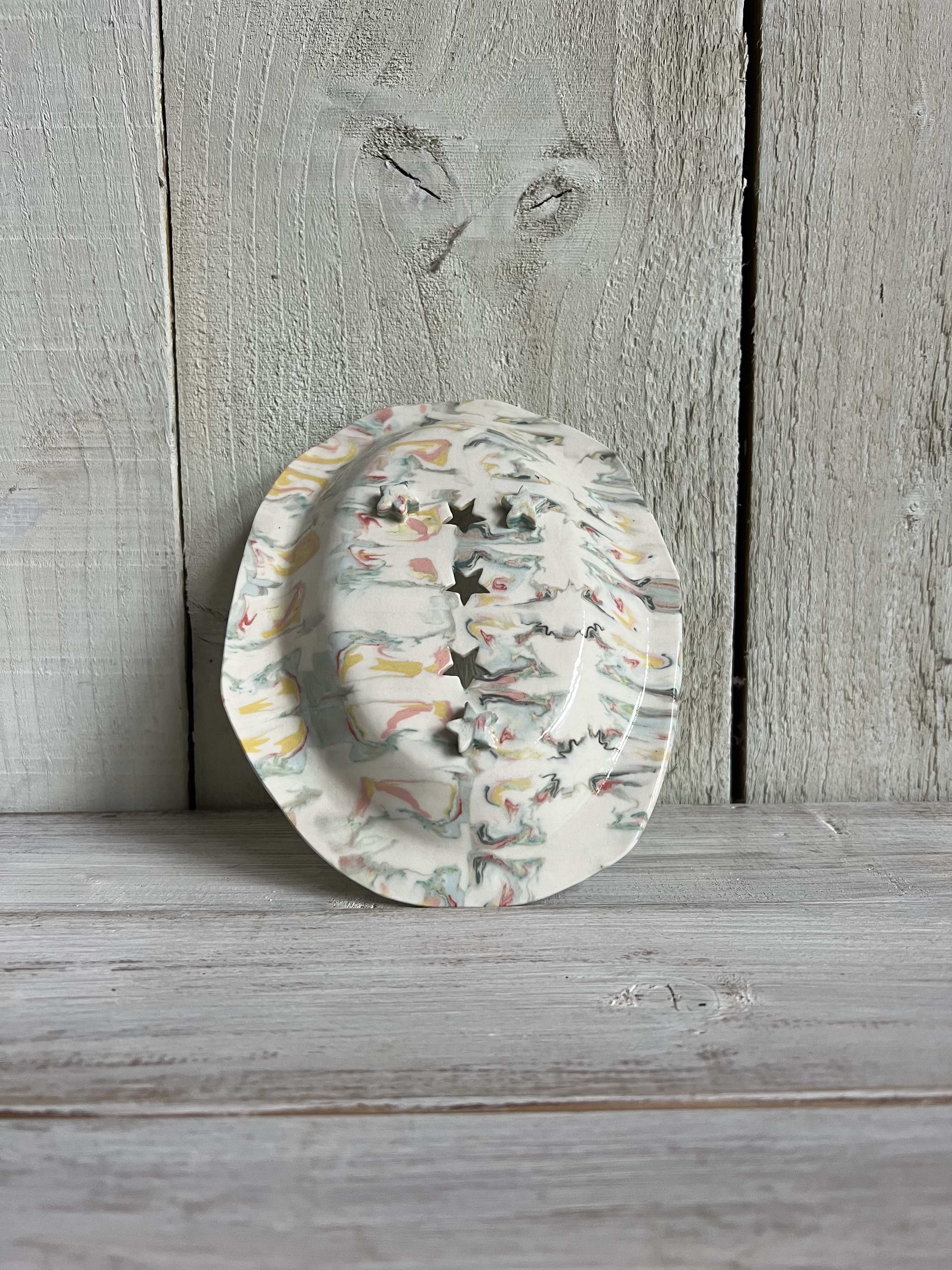 Pastel marbled handmade ceramic trinket dish with a scalloped edge and decorative star-shaped cut-outs standing on a rustic wooden shelf.