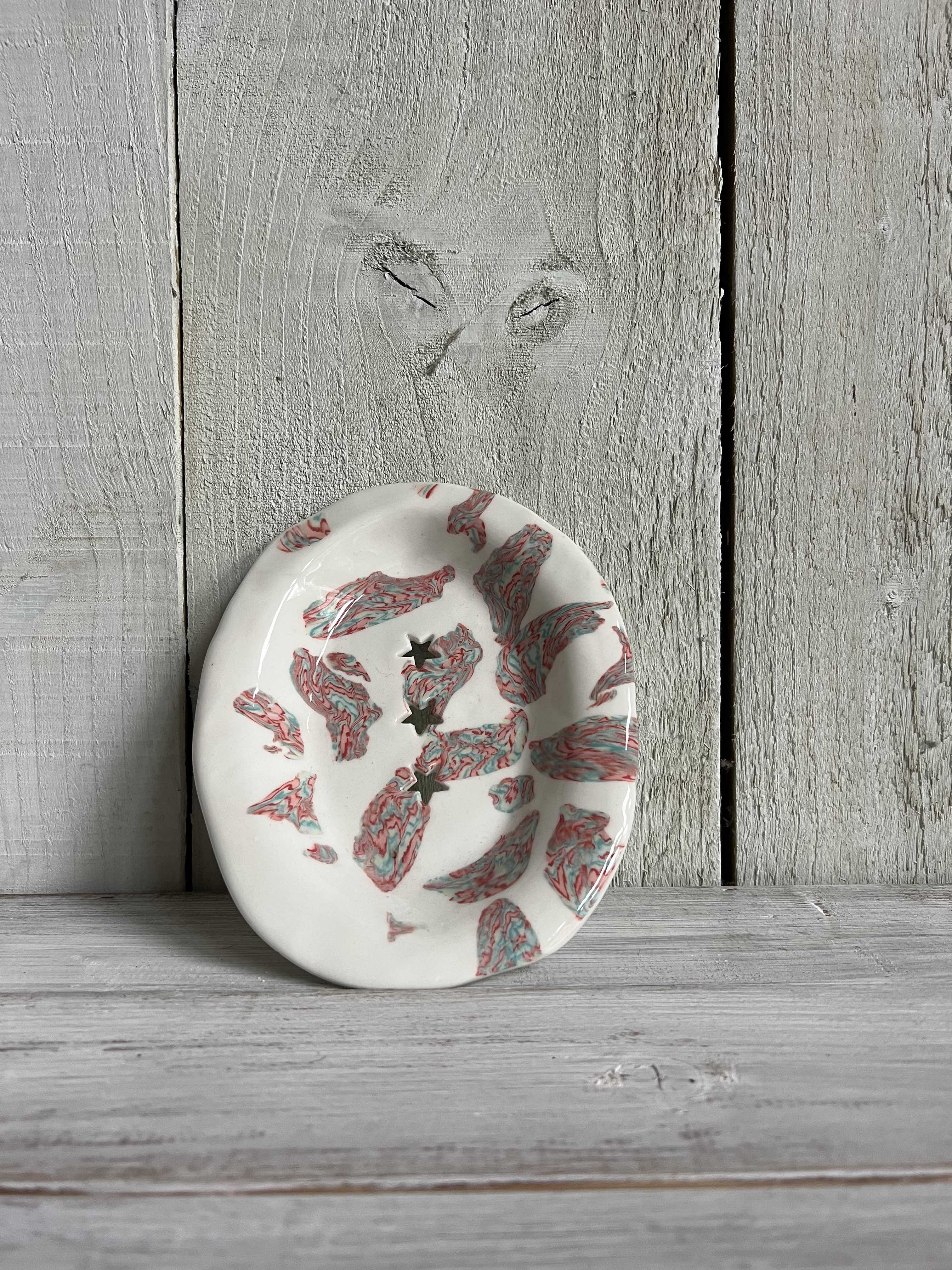 Handmade oval ceramic trinket dish with a white glaze, pink leaf pattern, and three star-shaped cut-outs displayed on a rustic wooden shelf.